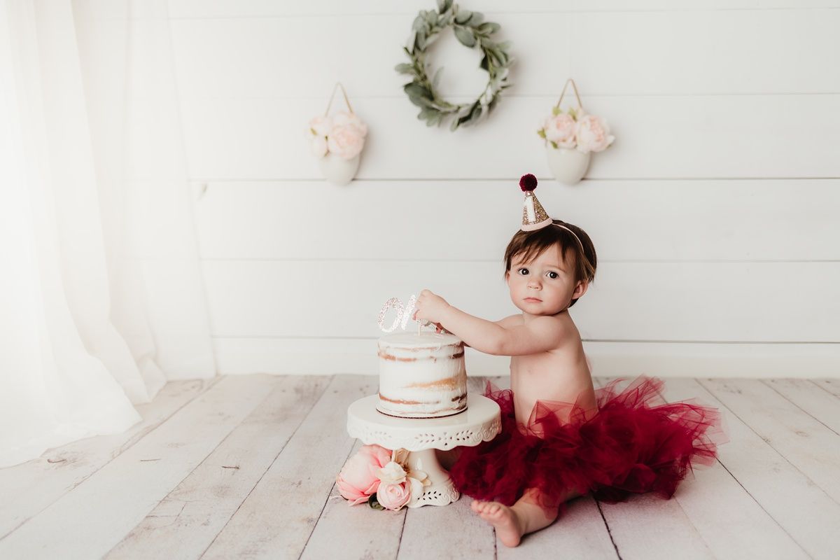 A small cake smash photo setup with a white cake, red tulle tutu, and rustic decorations against white shiplap.