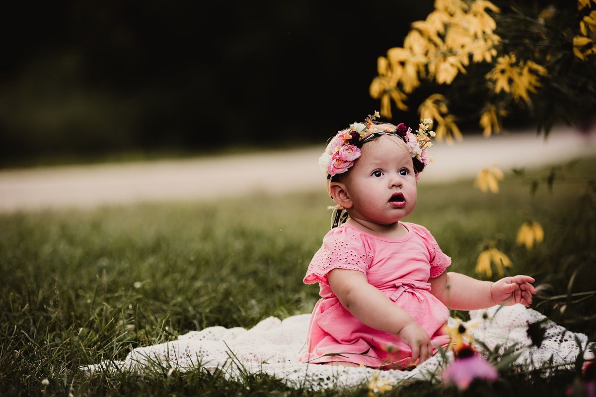 Baby in pink dress with flower crown sits on white blanket outdoors near autumn foliage.