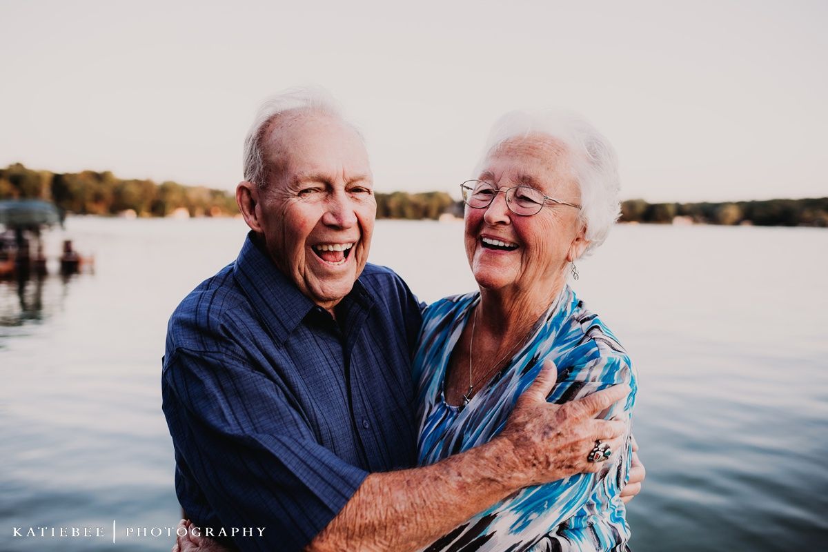 Elderly couple laughing together by a lake during sunset in a heartwarming outdoor portrait.