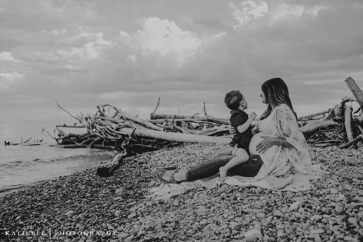 A black and white photo of a sweet moment between loved ones on a rocky beach with driftwood.