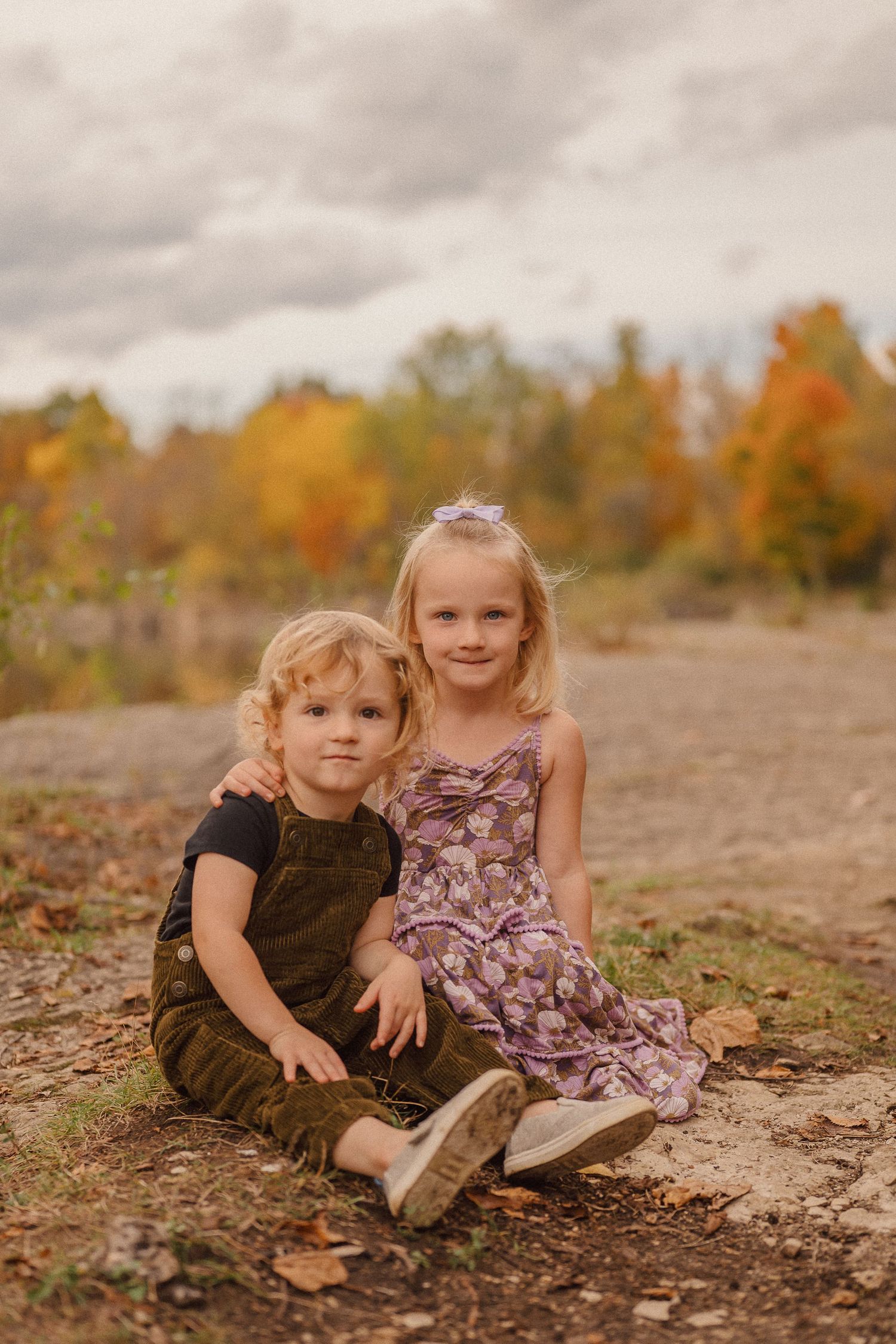 Two young children sit together on rocky ground surrounded by autumn foliage in a warm-toned portrait setting.
