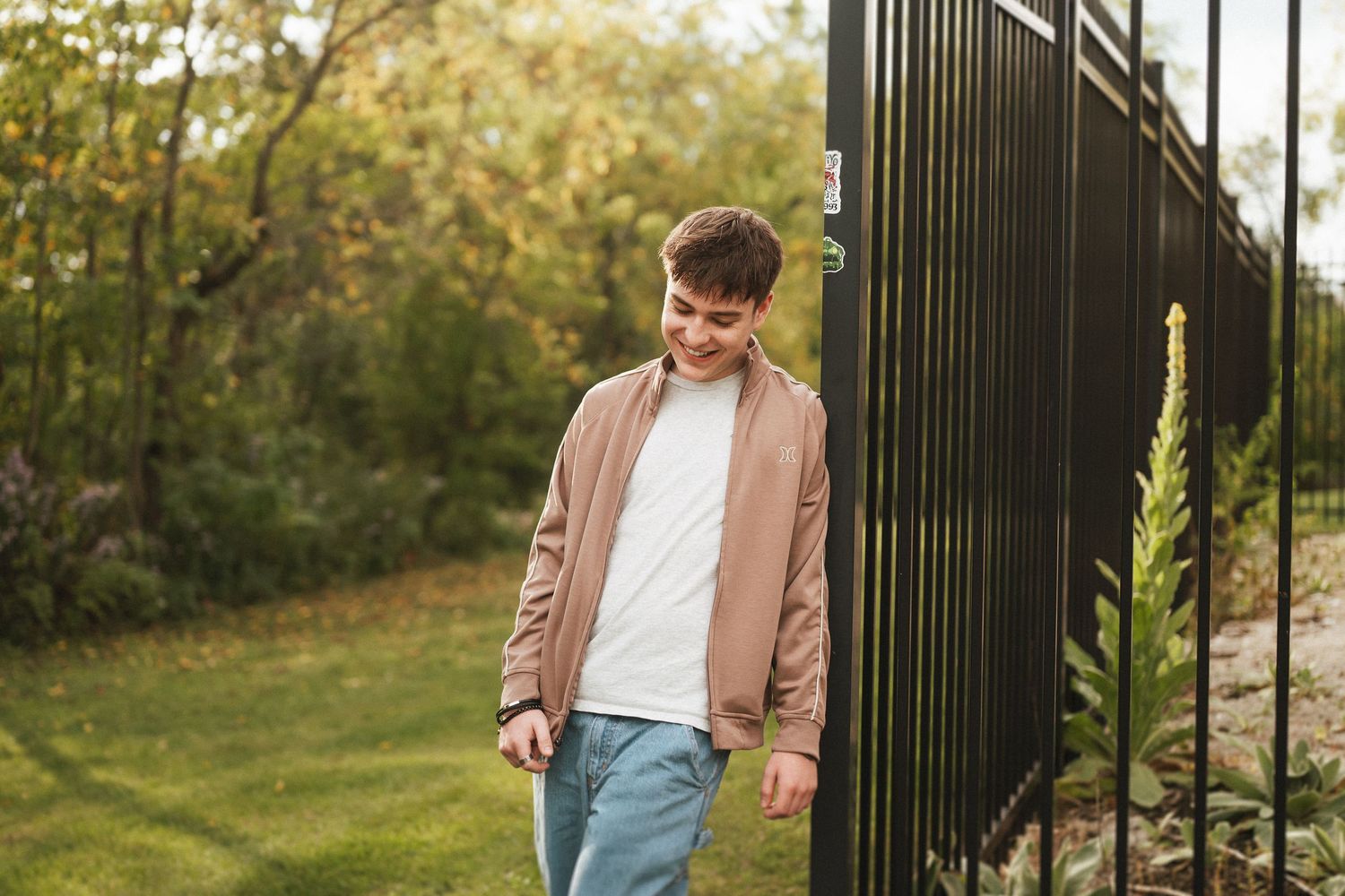 Person in a brown jacket and jeans walks casually through an autumn garden setting with iron gates and greenery.