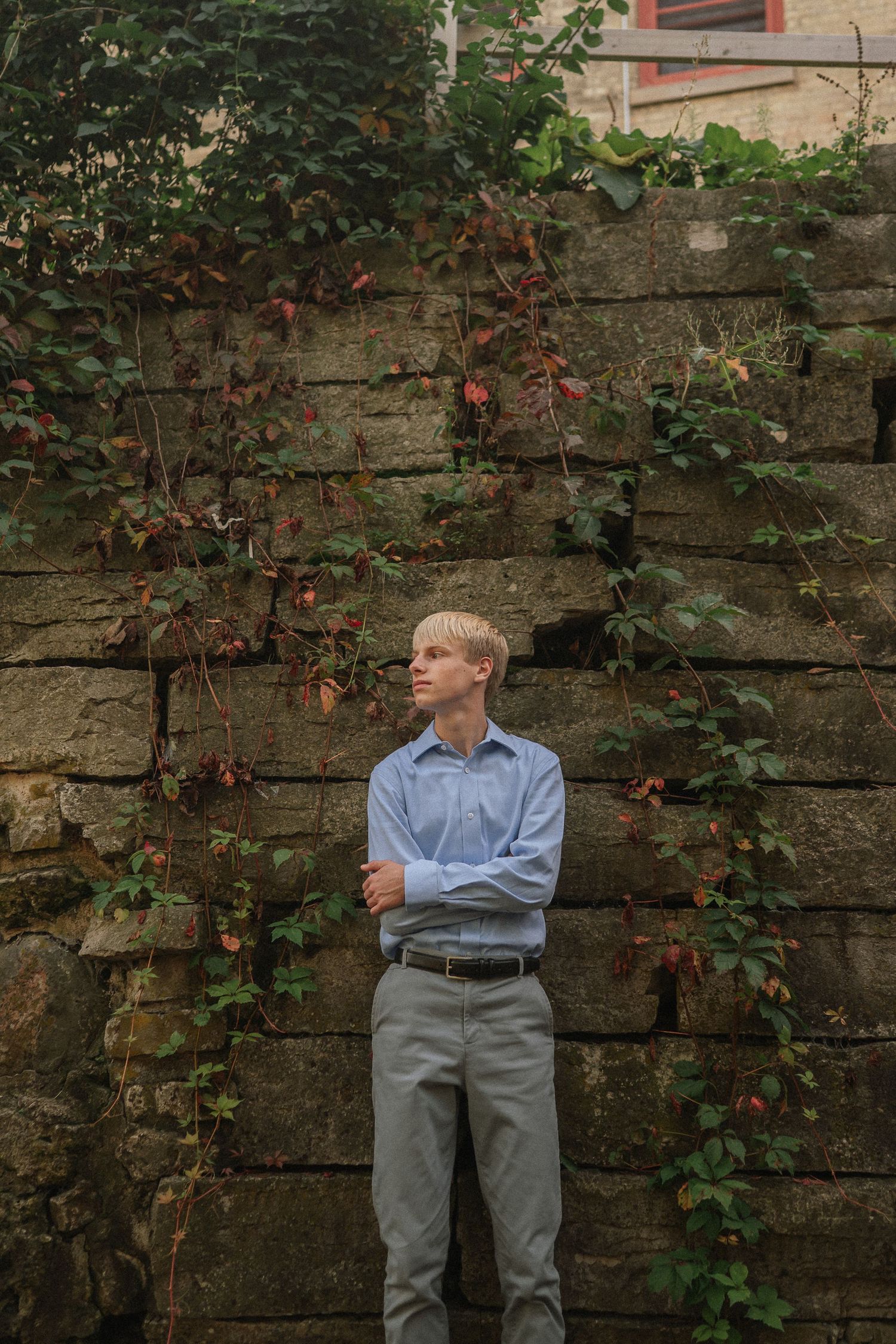 A figure in a light blue shirt and gray pants poses against a stone wall with climbing vines in a garden setting.