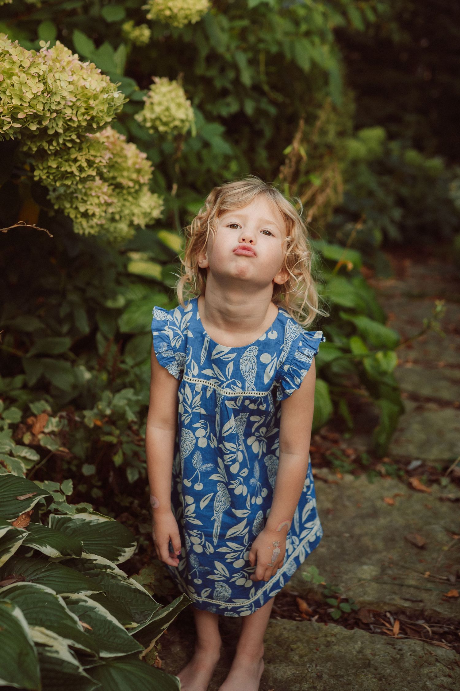 Young child in blue floral dress making silly faces in a garden setting.