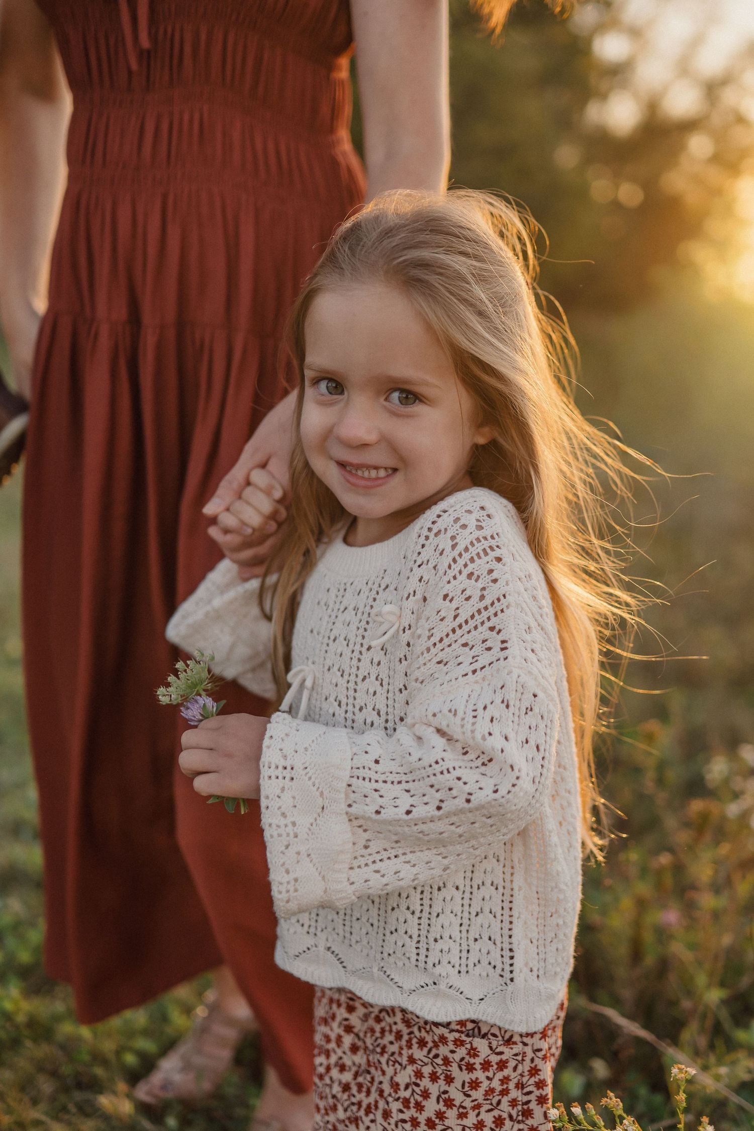 A young girl in a white sweater and patterned skirt twirls playfully in a sunlit meadow.