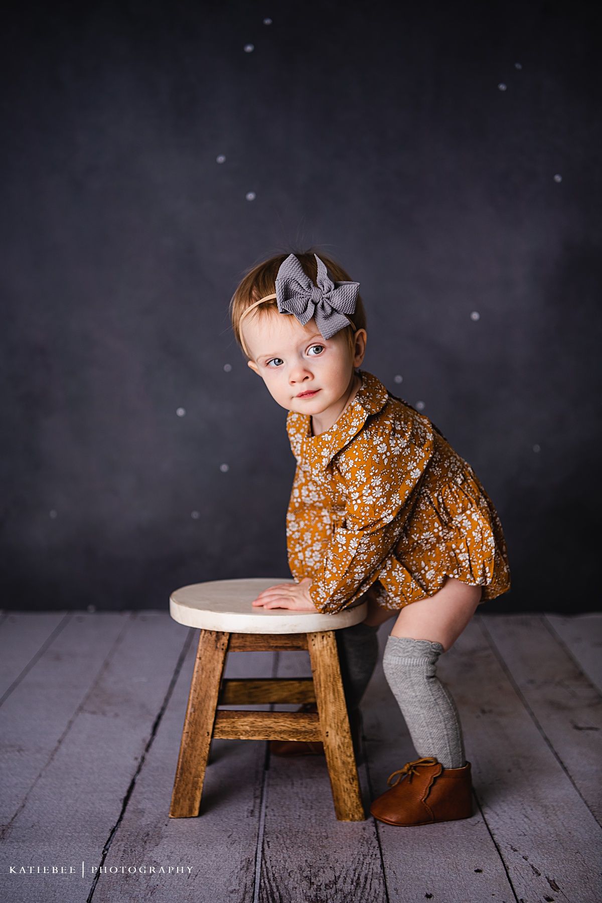 Child in mustard floral outfit and gray bow headband leaning on wooden stool.