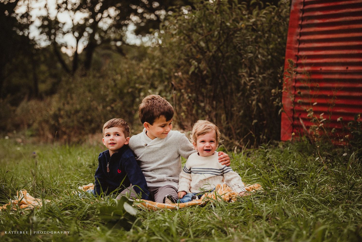Three siblings sitting together in tall grass beside a red barn.