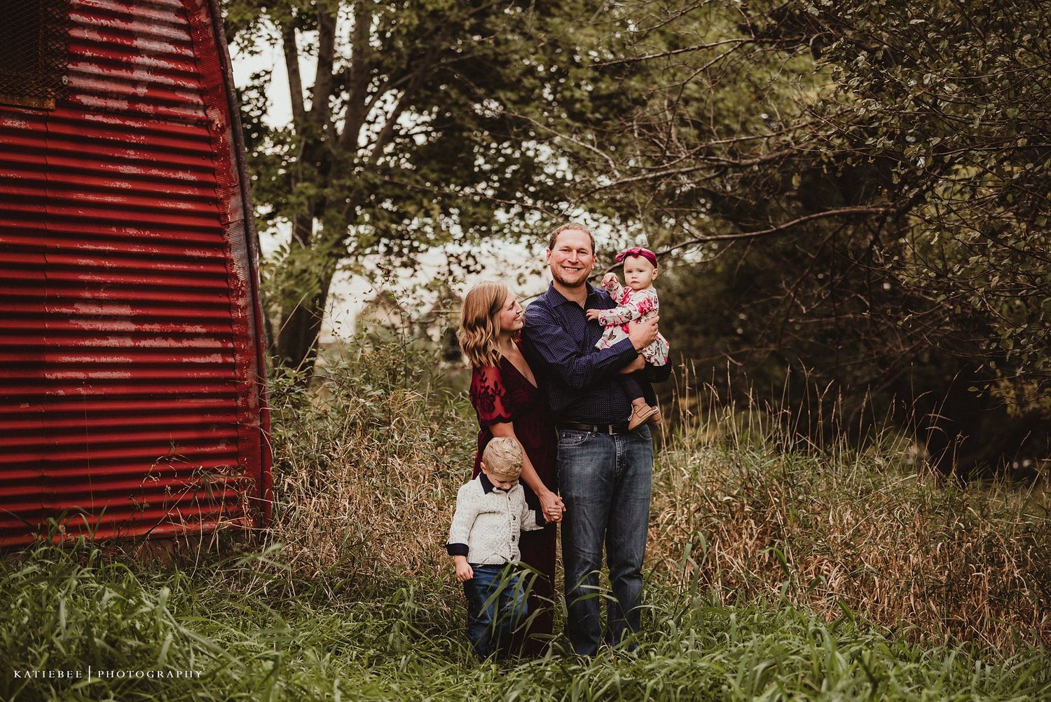 A family portrait in front of a red barn amongst tall grass and trees.