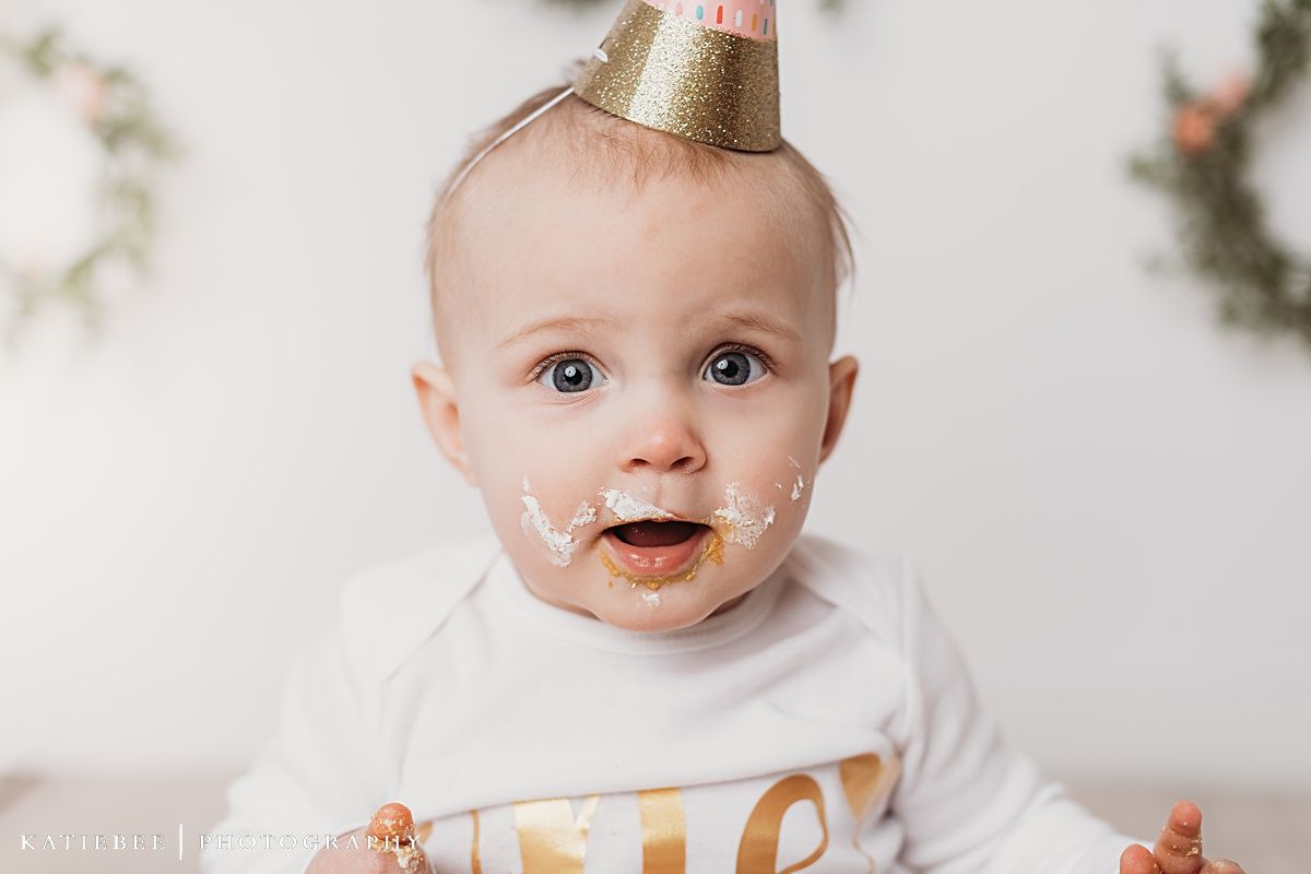 Baby in white shirt with gold and pink party hat smiling with cake frosting on face.