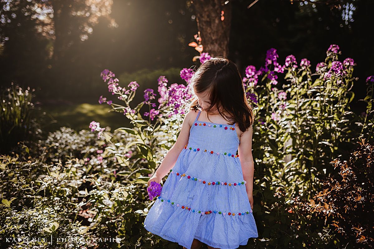 A child in a blue dress explores a garden with blooming purple flowers at sunset.