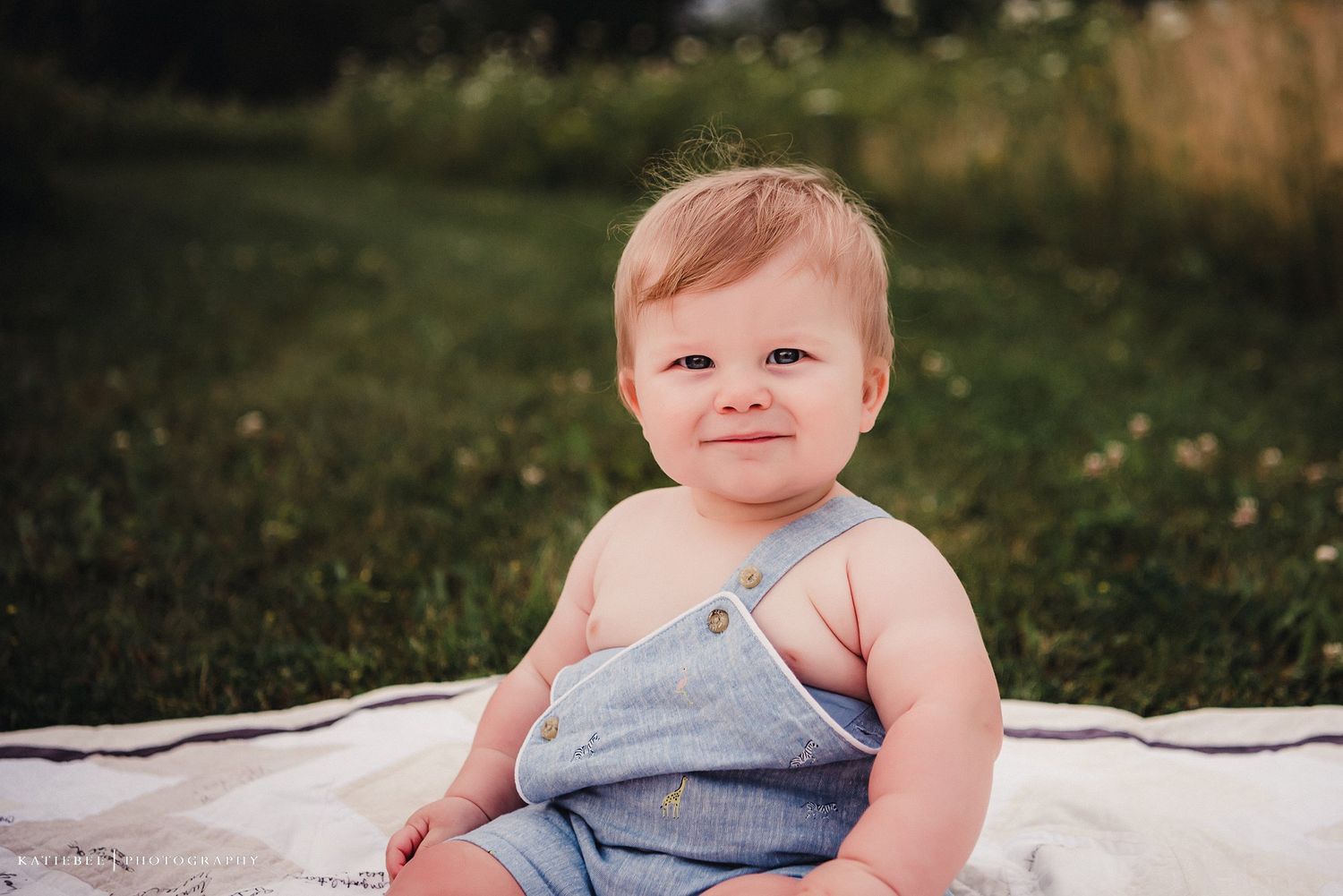A smiling baby in blue overalls sits on a white blanket outdoors.