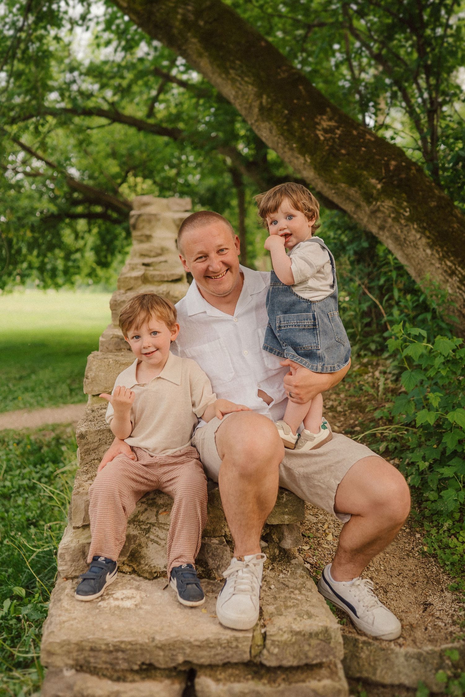 A warm family moment captured on stone steps under a large tree in a lush garden setting.