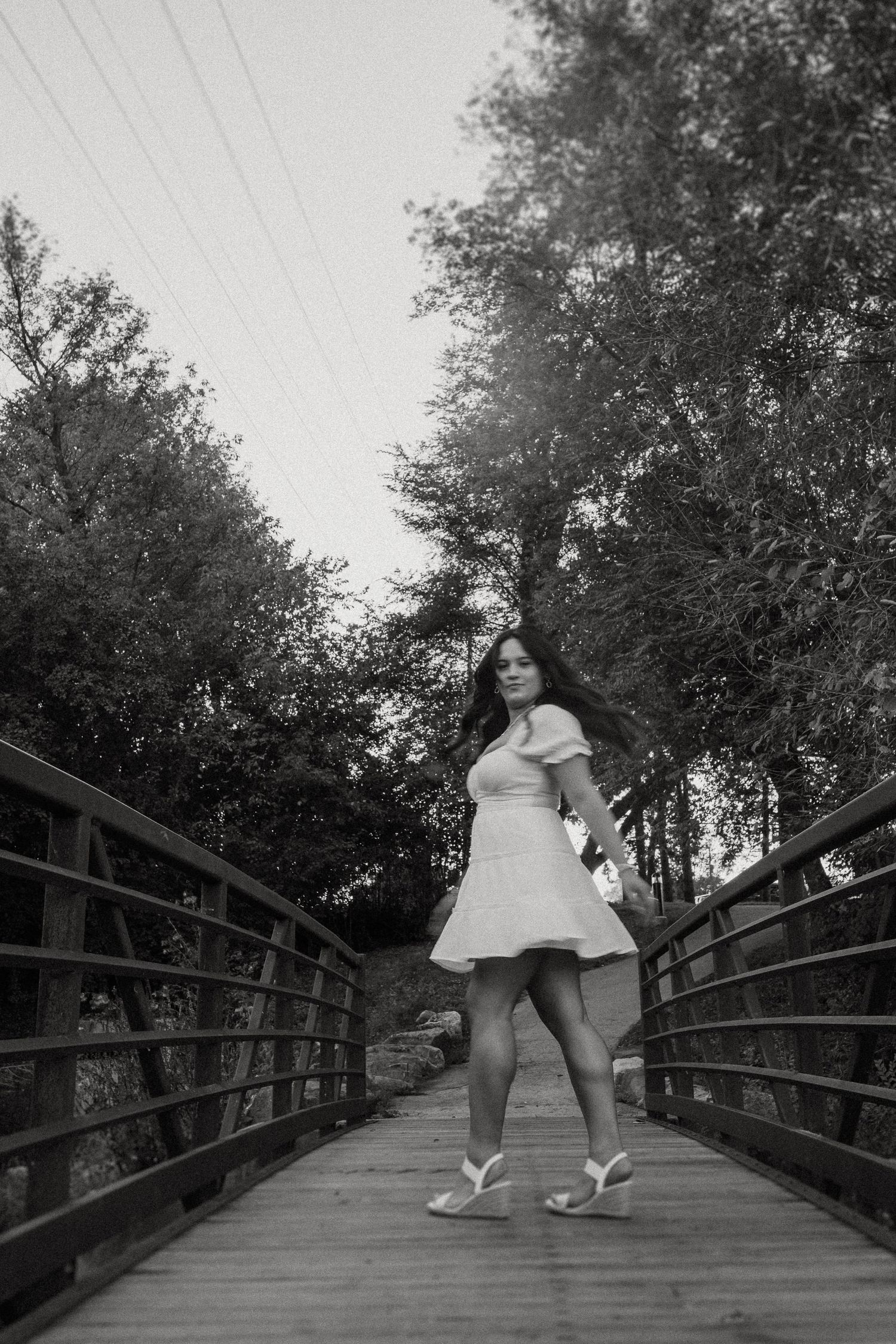 A black and white photo of a person in a white dress standing on a wooden footbridge.