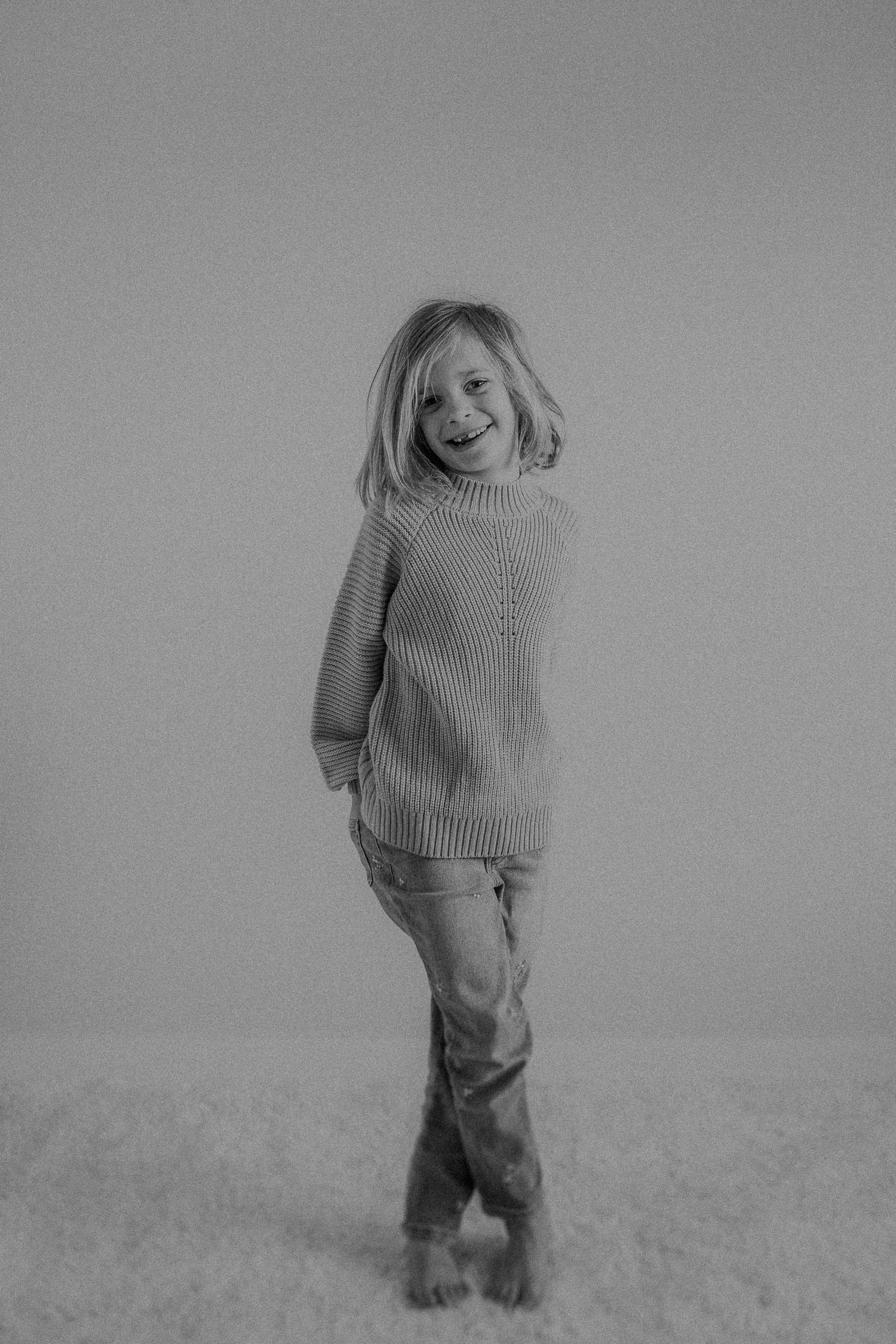 Black and white portrait showing playful expression while wearing a cozy sweater and jeans on light background.