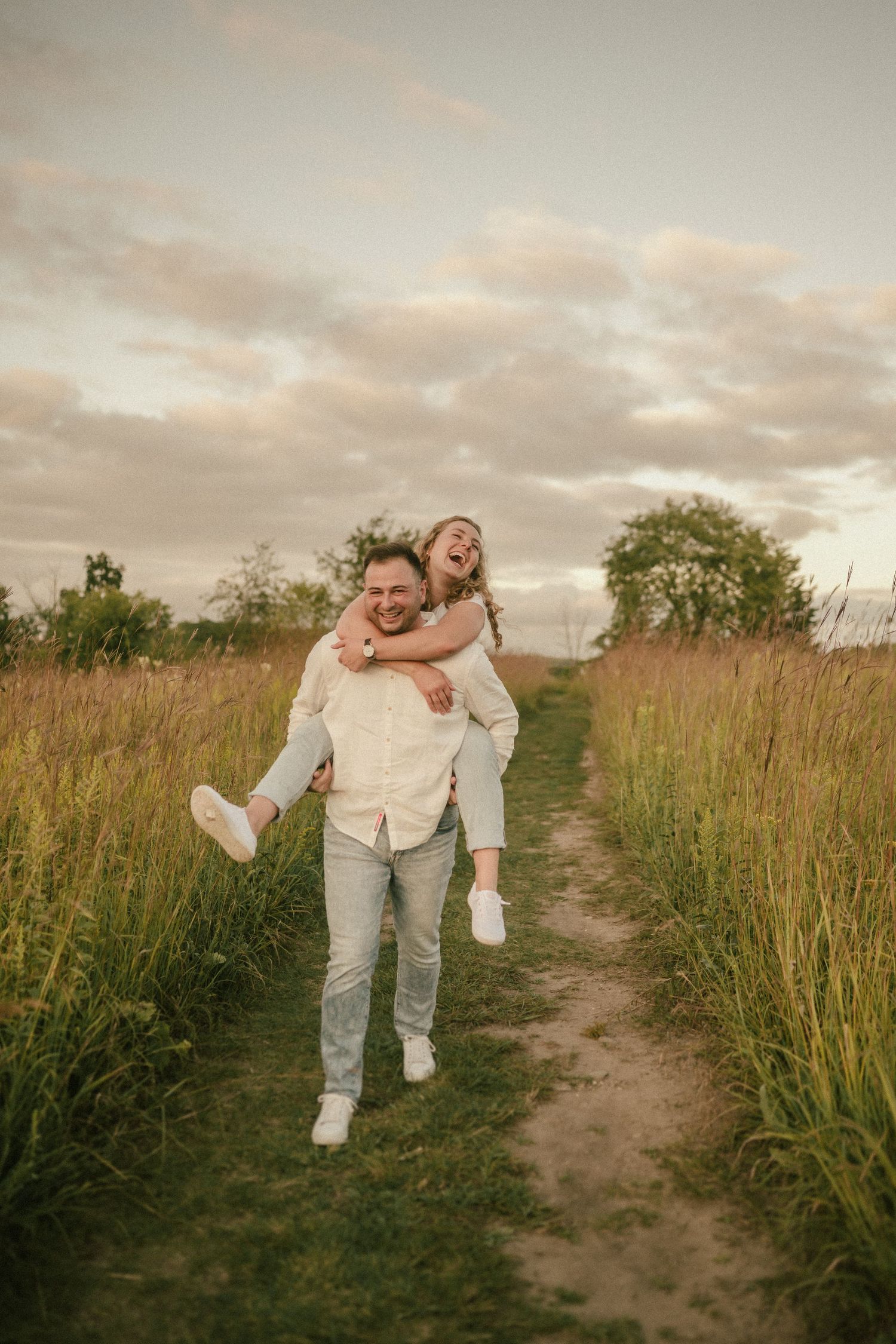 Playful piggyback ride along nature trail surrounded by tall grass at sunset.