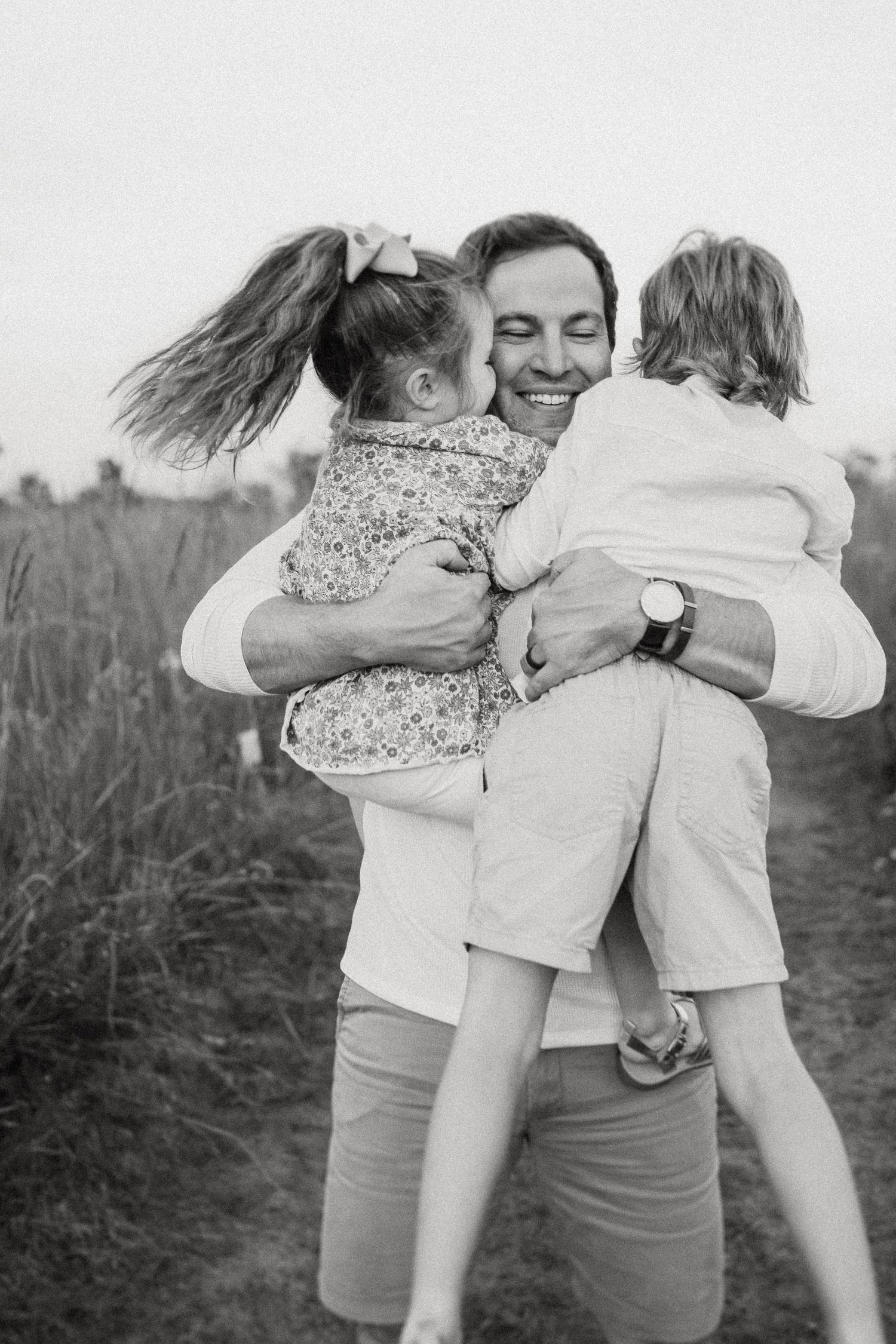 A joyful embrace captured in black and white with two children being held in a grassy field at sunset.