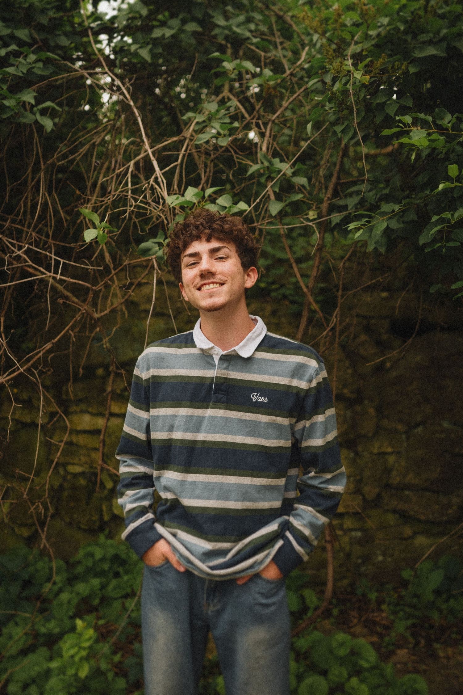 A person wearing a striped polo sweater stands smiling in front of a garden wall with green foliage.