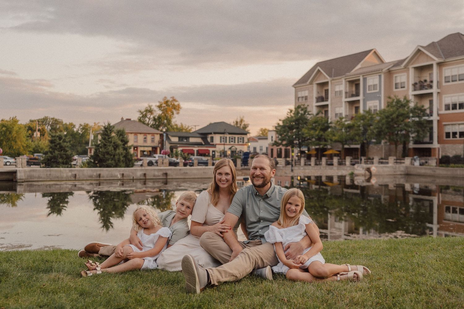 Family relaxes together on grassy hill during golden hour sunset near waterfront residential buildings.