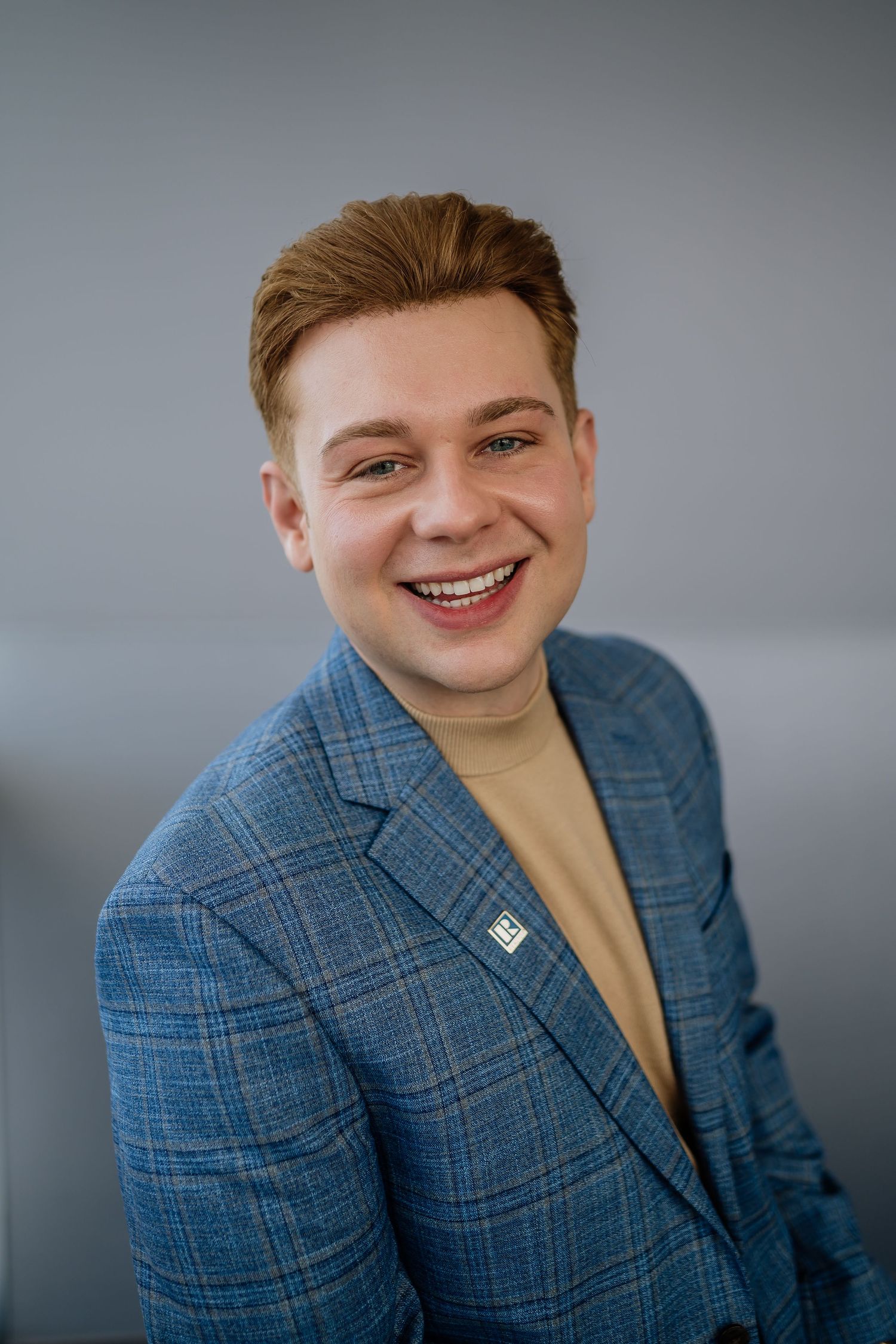 Professional headshot of someone wearing a blue plaid blazer and tan turtleneck with a warm smile against grey background.