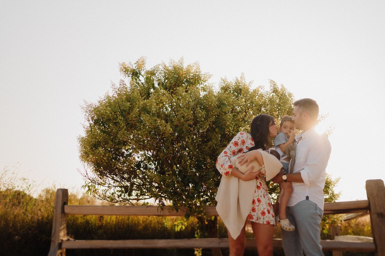 Warm sunset light illuminates family moment by rustic fence and greenery.