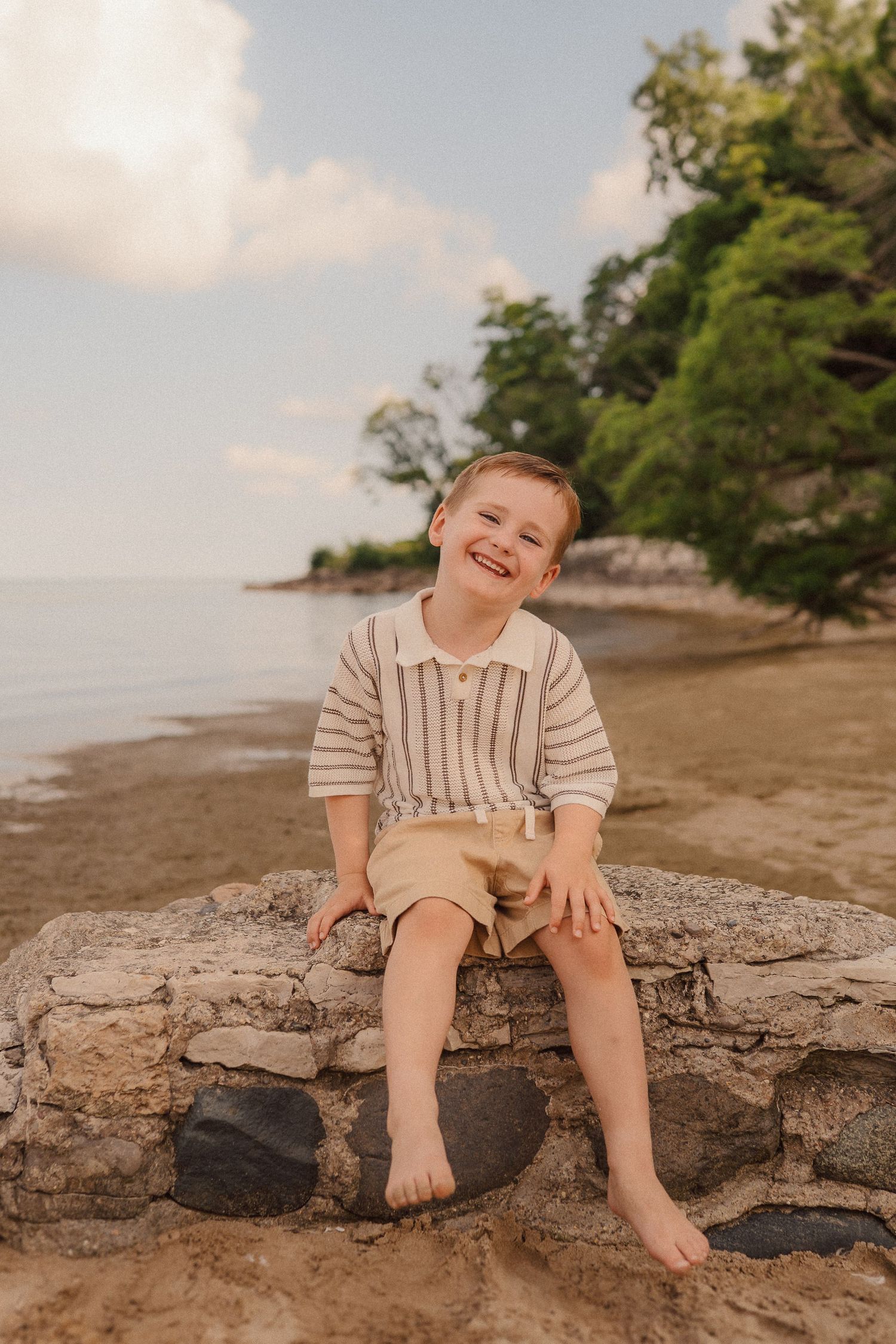 Child sitting barefoot on rocky beach wall wearing striped sweater and khaki shorts at sunset.