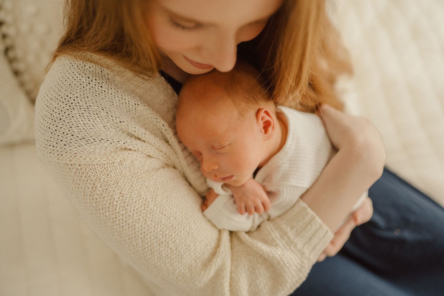 A tender moment as a sleeping newborn is cradled against a beige knit sweater in warm natural light.