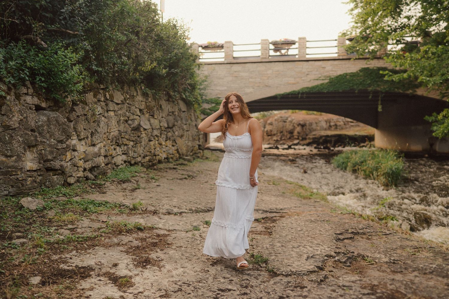 A person in a white wedding dress stands on a dry riverbed beneath a stone bridge during golden hour.