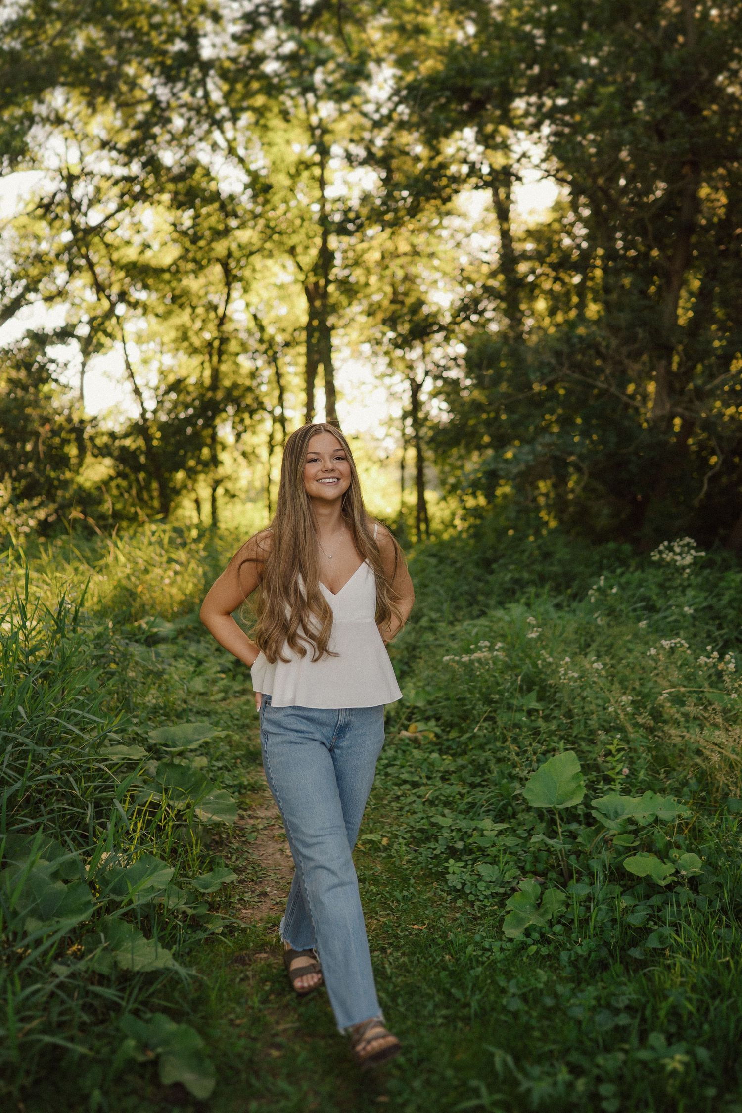 A woman in a white tank top and jeans poses in a sunlit forest path during golden hour.
