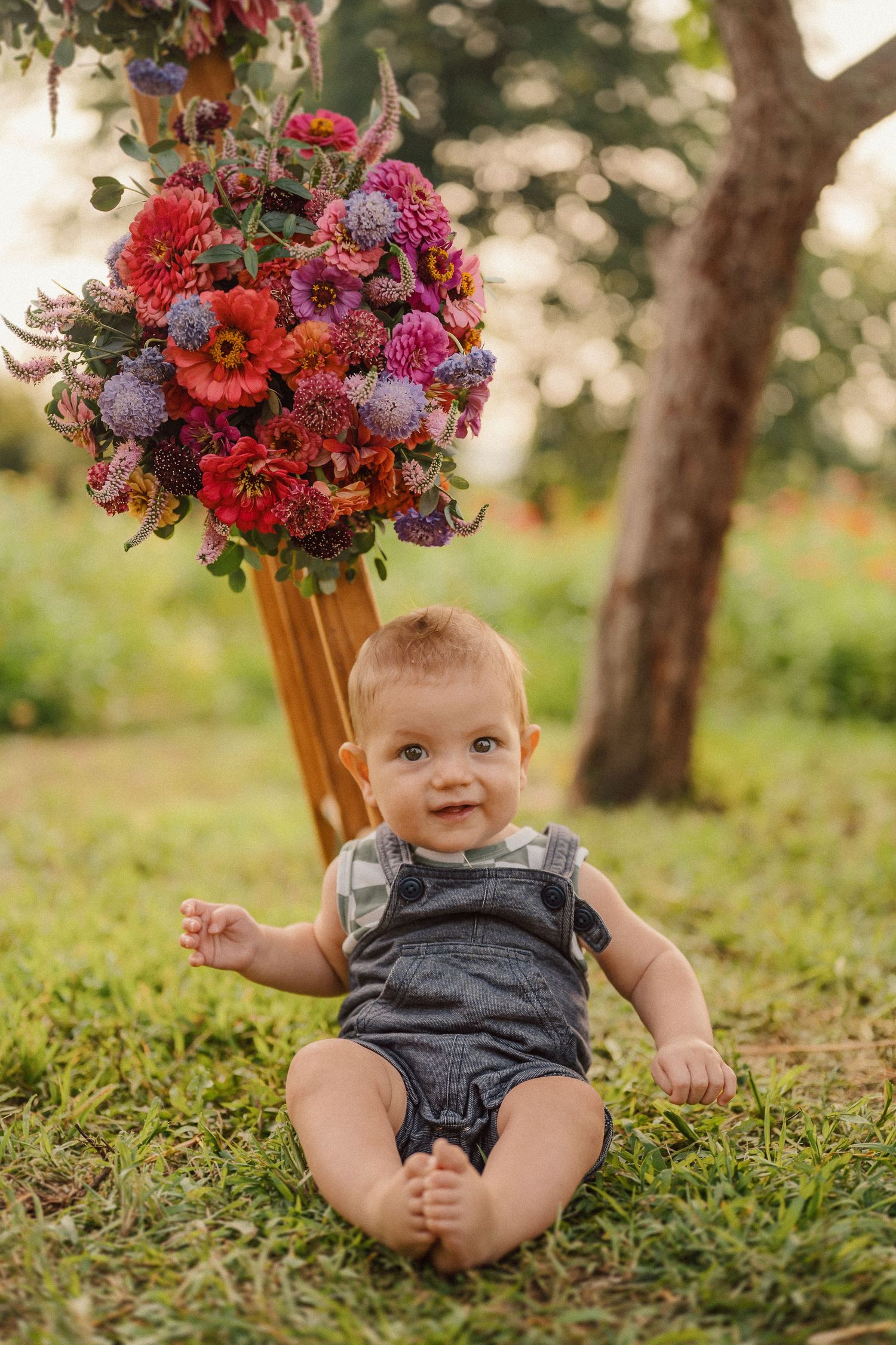 A baby in grey overalls sits on grass beneath a vibrant red and purple floral arrangement.