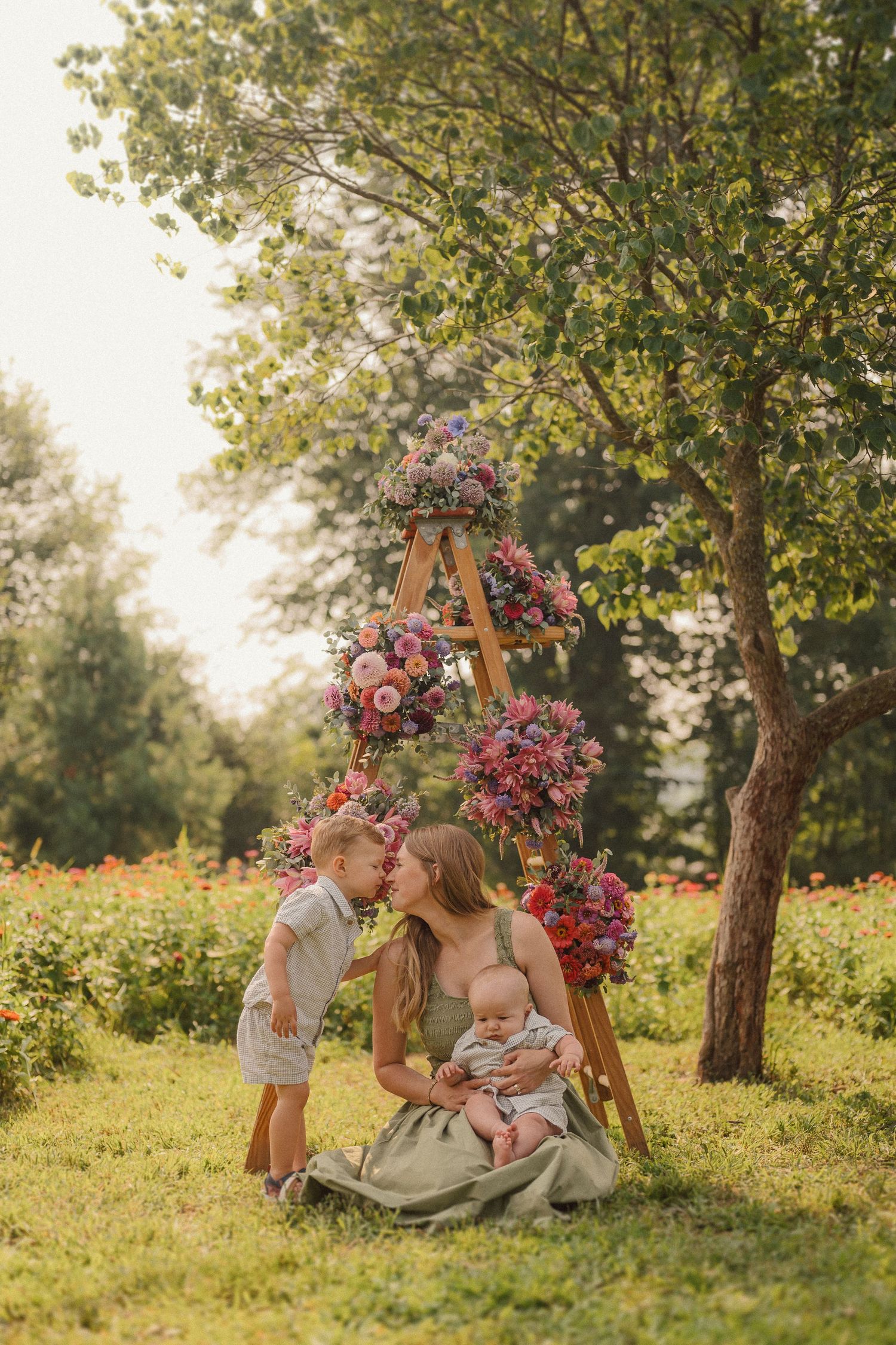 Family sits together on grass under wooden archway decorated with pink flowers during golden sunset in park.