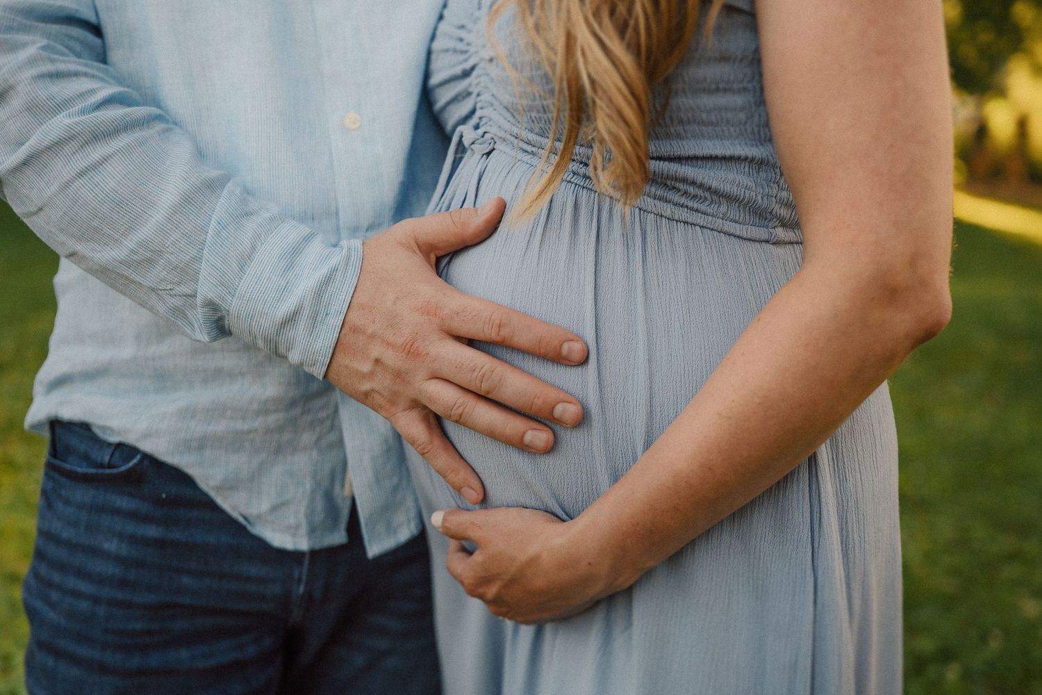 Close-up photo of pregnant belly in light blue shirt with hands gently resting on the bump during outdoor photoshoot.