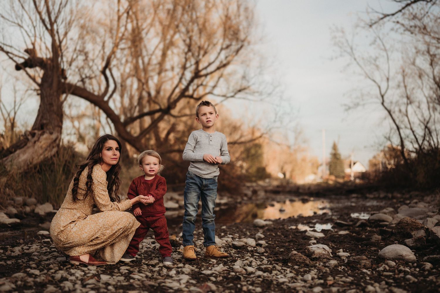 Family enjoying time together on rocky ground near a stream during sunset with autumn trees behind them.