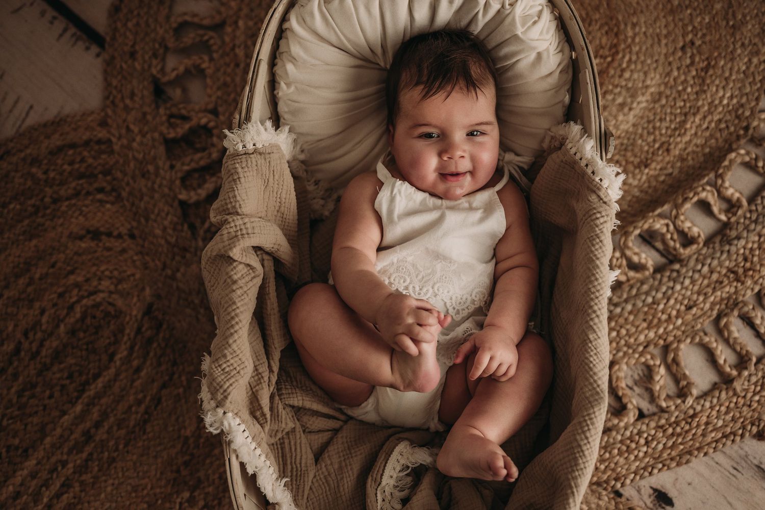 Baby in white outfit reclines in vintage-style beige bassinet on jute rug.