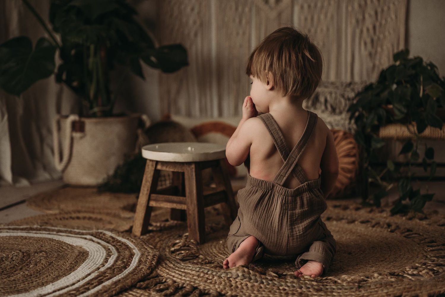 Small child in brown romper sitting beside wooden stool on textured rug with macrame wall decor.