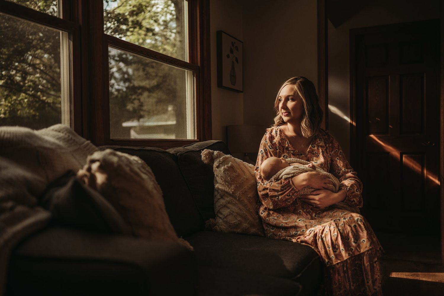 Mother in floral dress holds sleeping newborn on couch by window with natural lighting.