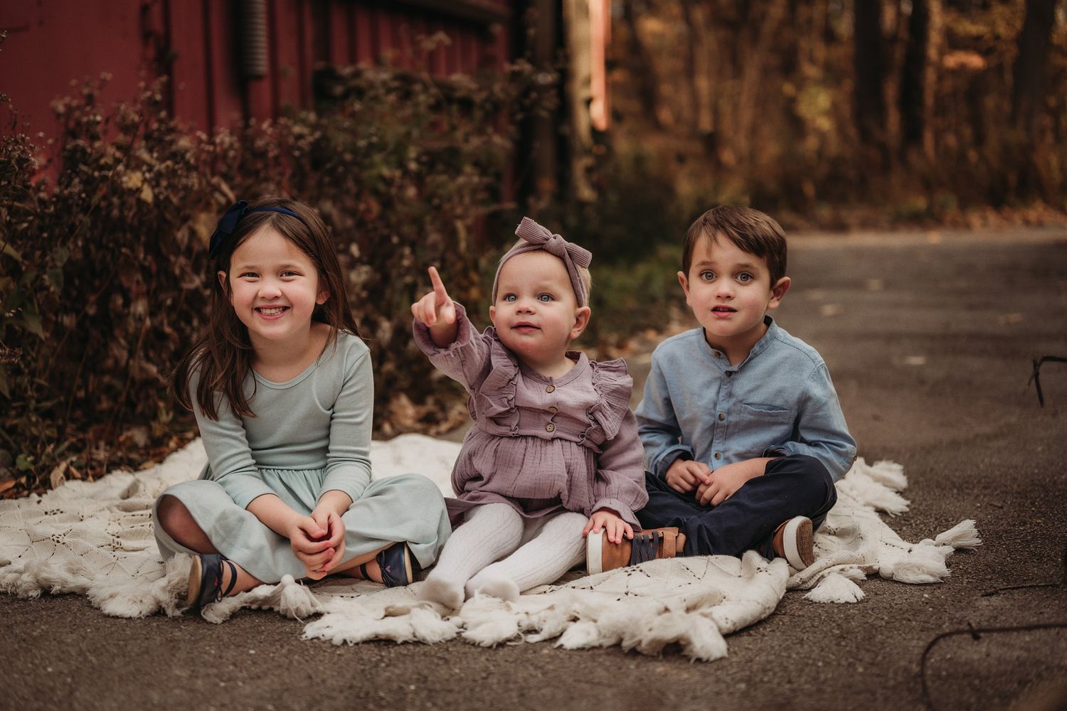 Three children sit on white blankets on a path near autumn foliage.