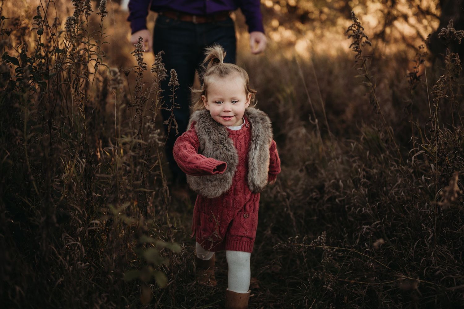 A toddler in a red sweater and fur vest playing outdoors in tall grass.
