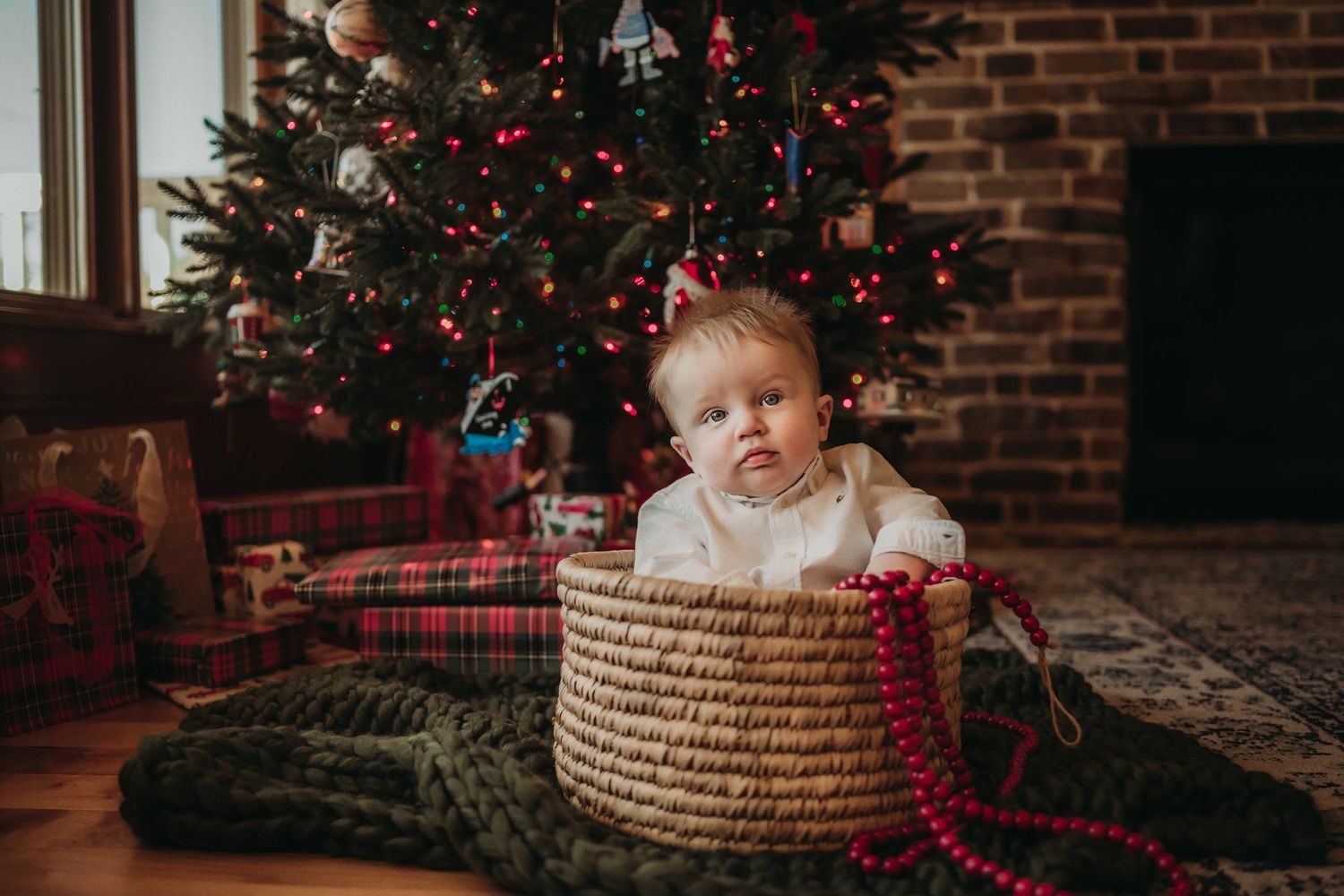A cozy Christmas scene with a woven basket and decorated tree on plaid blankets near a brick fireplace.