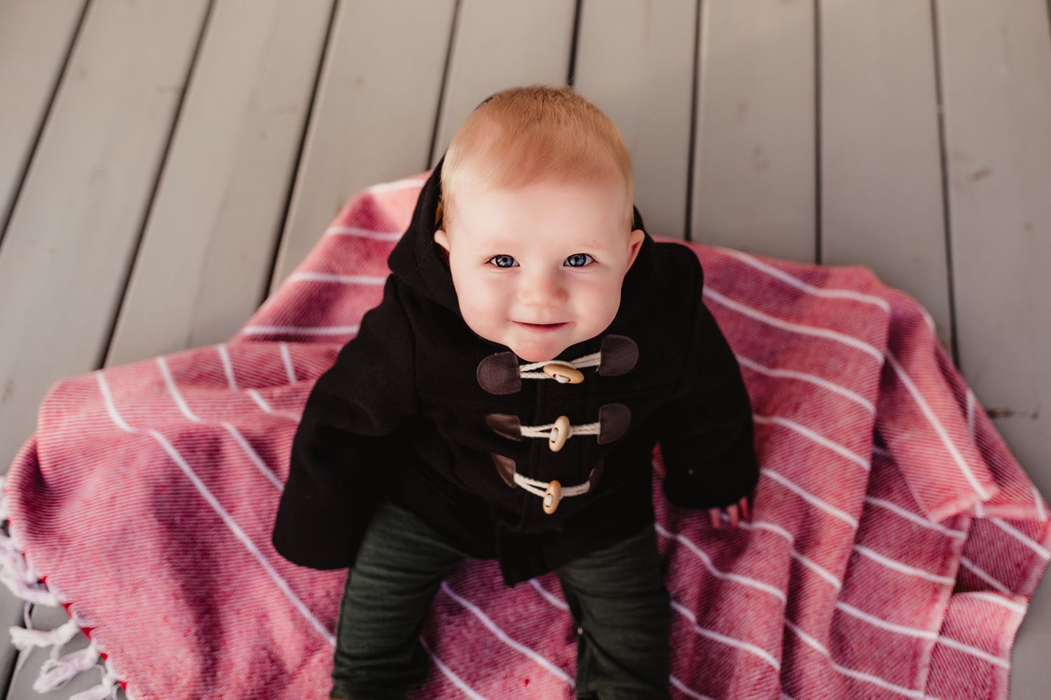Smiling baby wearing black toggle coat sitting on pink striped blanket.