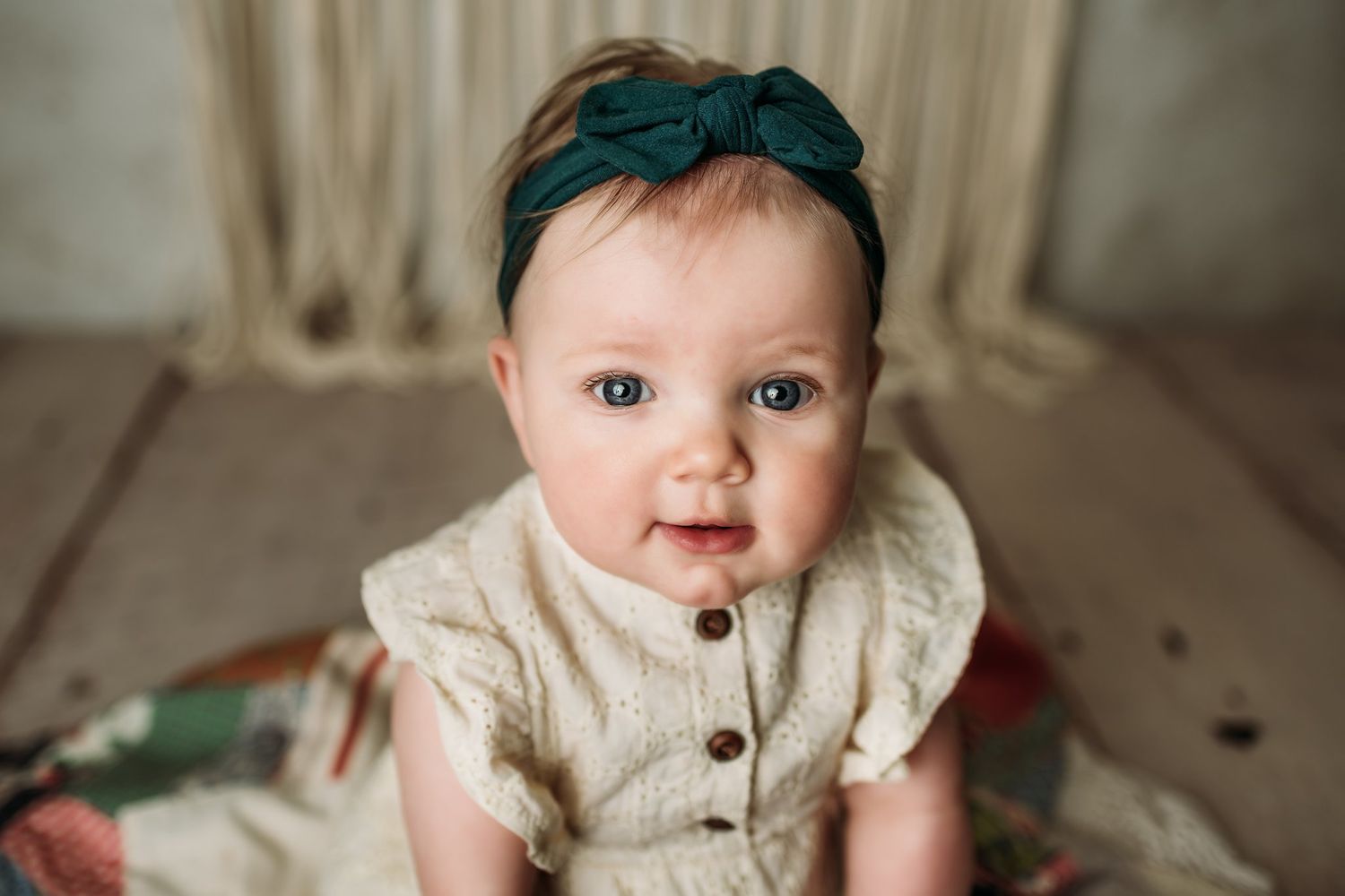 Close-up portrait of baby in cream dress with teal headband against neutral background.