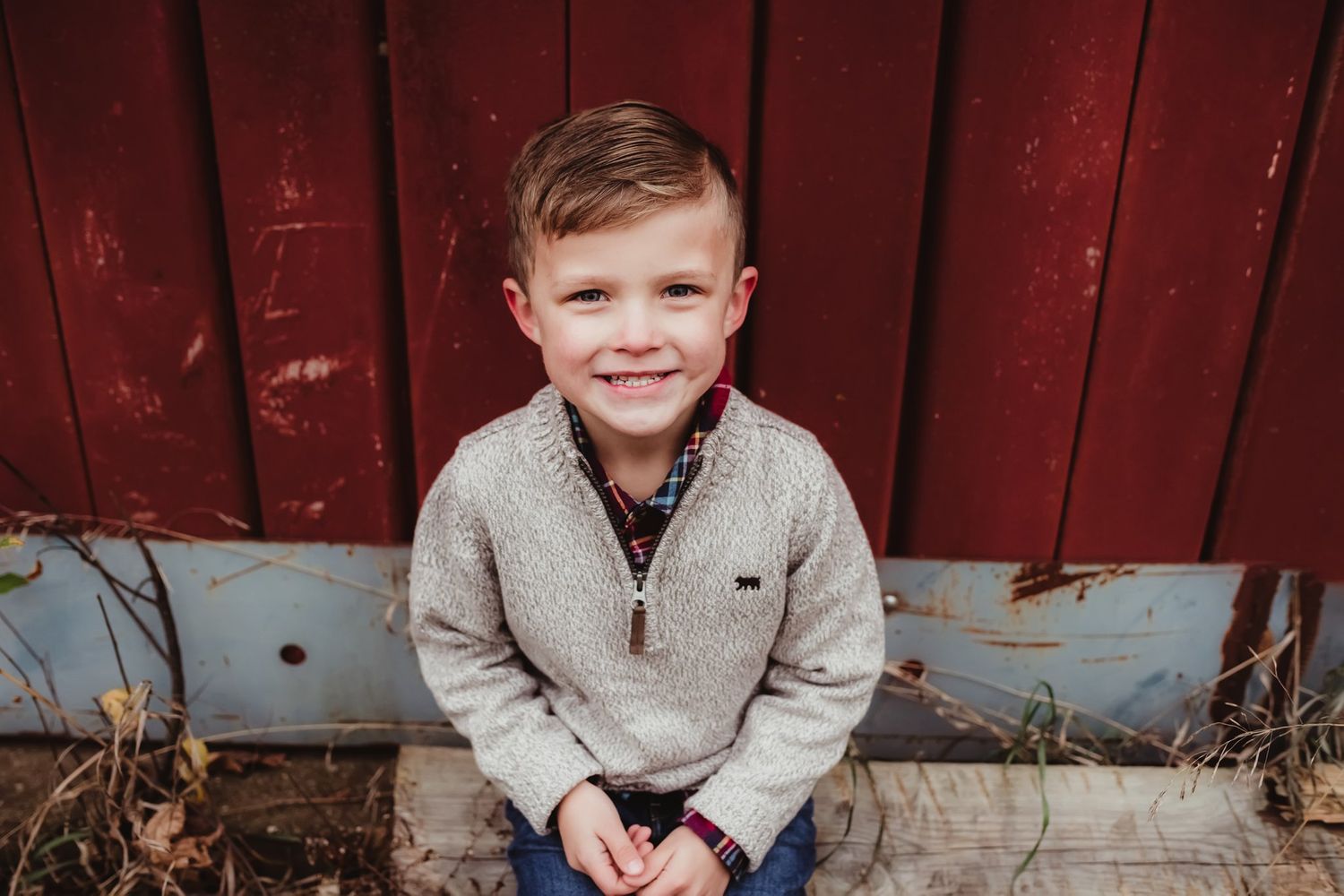 A child in a beige sweater sits by a red barn wall with a cheerful expression.