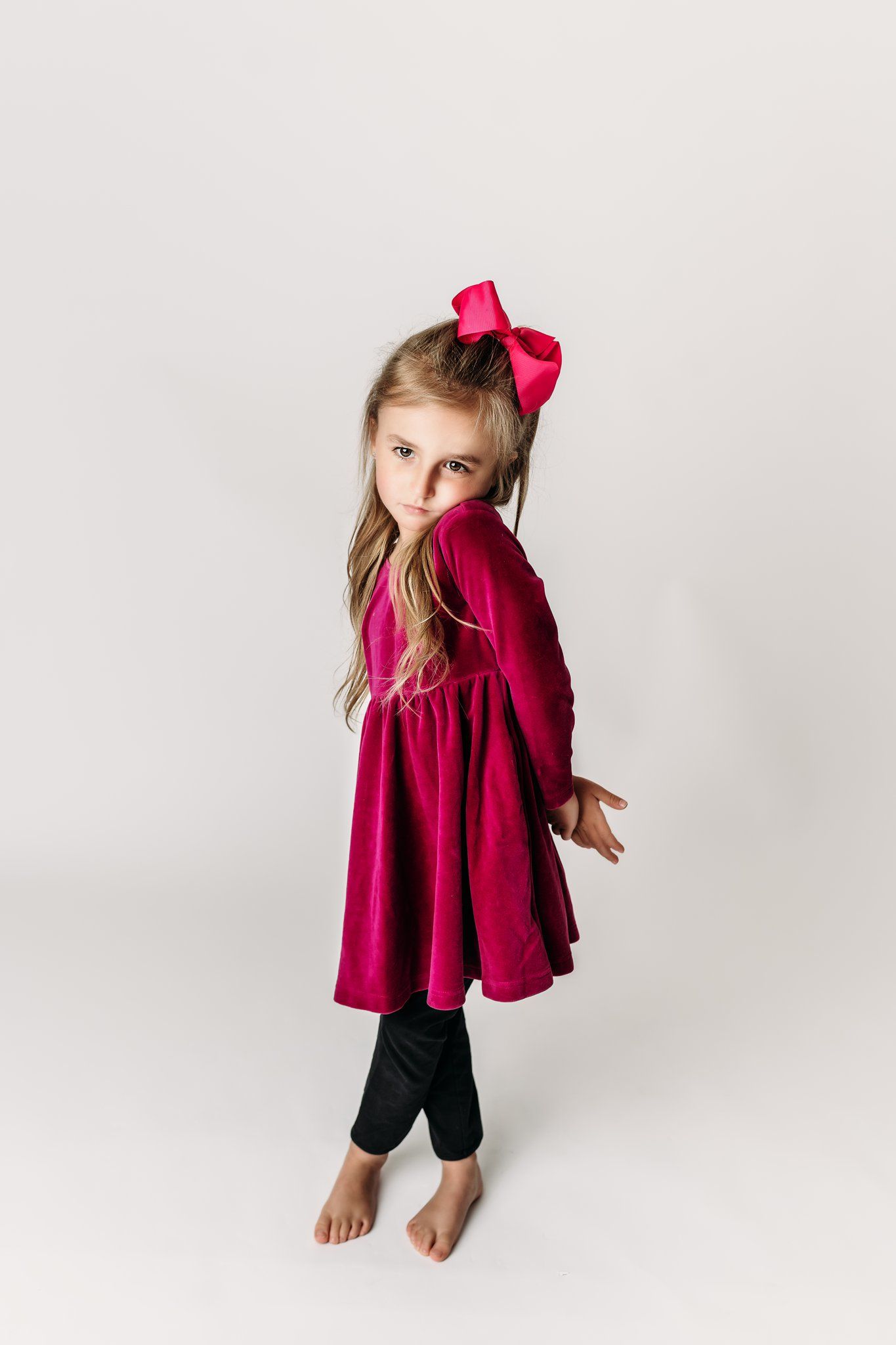 Child in burgundy dress and leggings strikes playful pose on white backdrop.