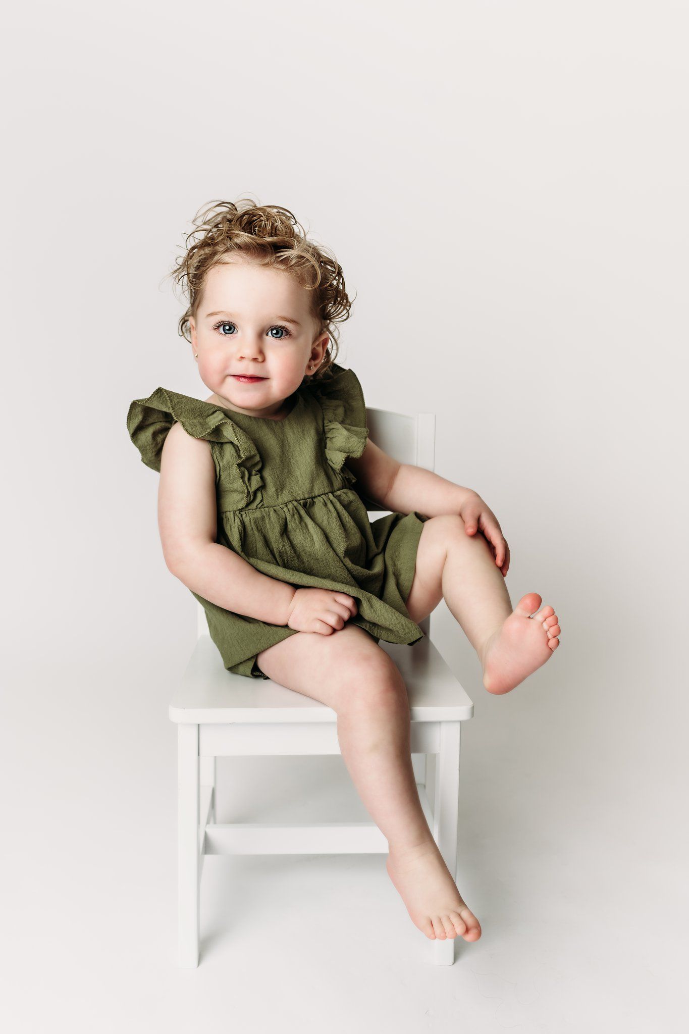 Child in olive green dress sits on white chair against neutral background.