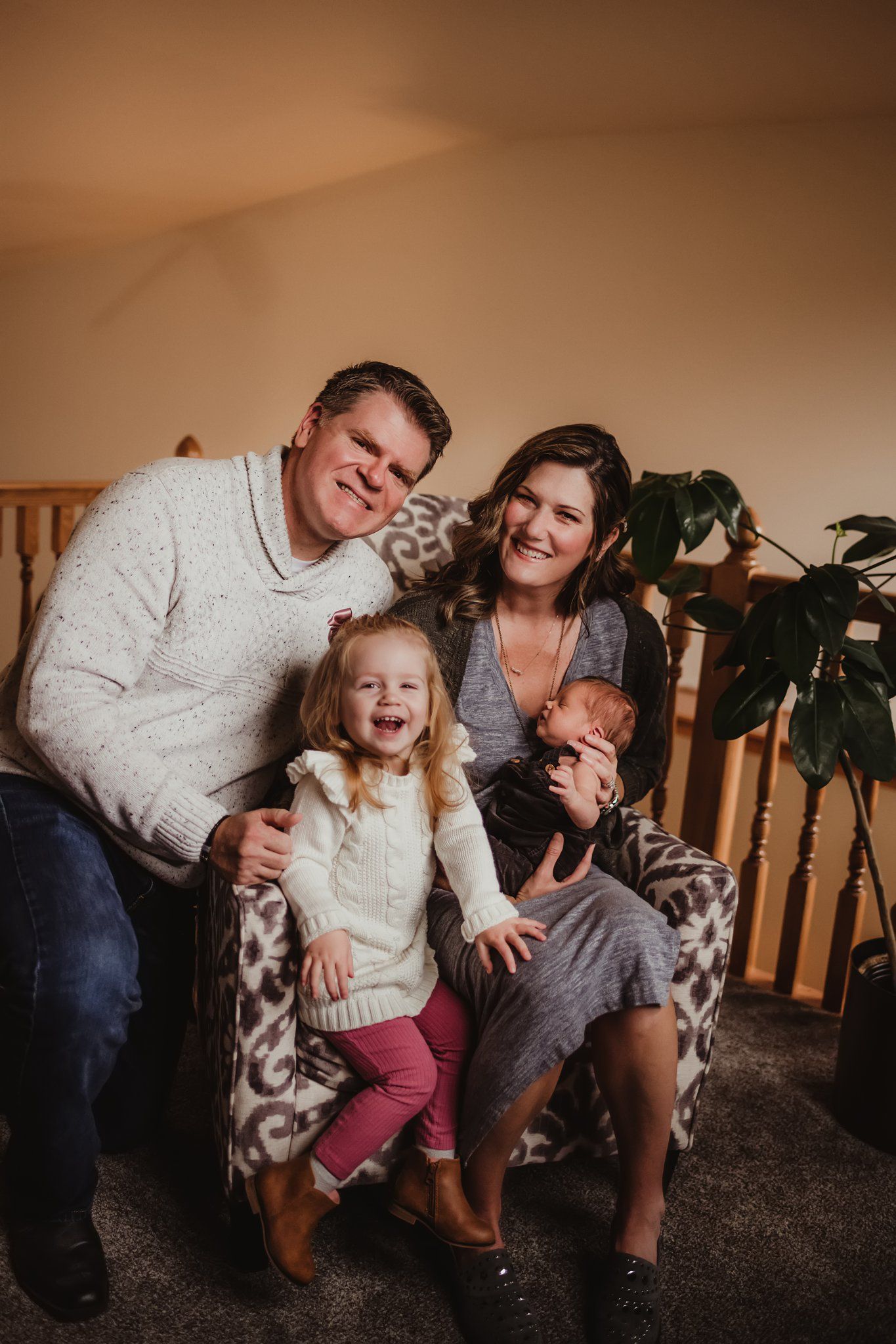 A family of four sits together on a patterned accent chair in front of wooden railings and a houseplant.