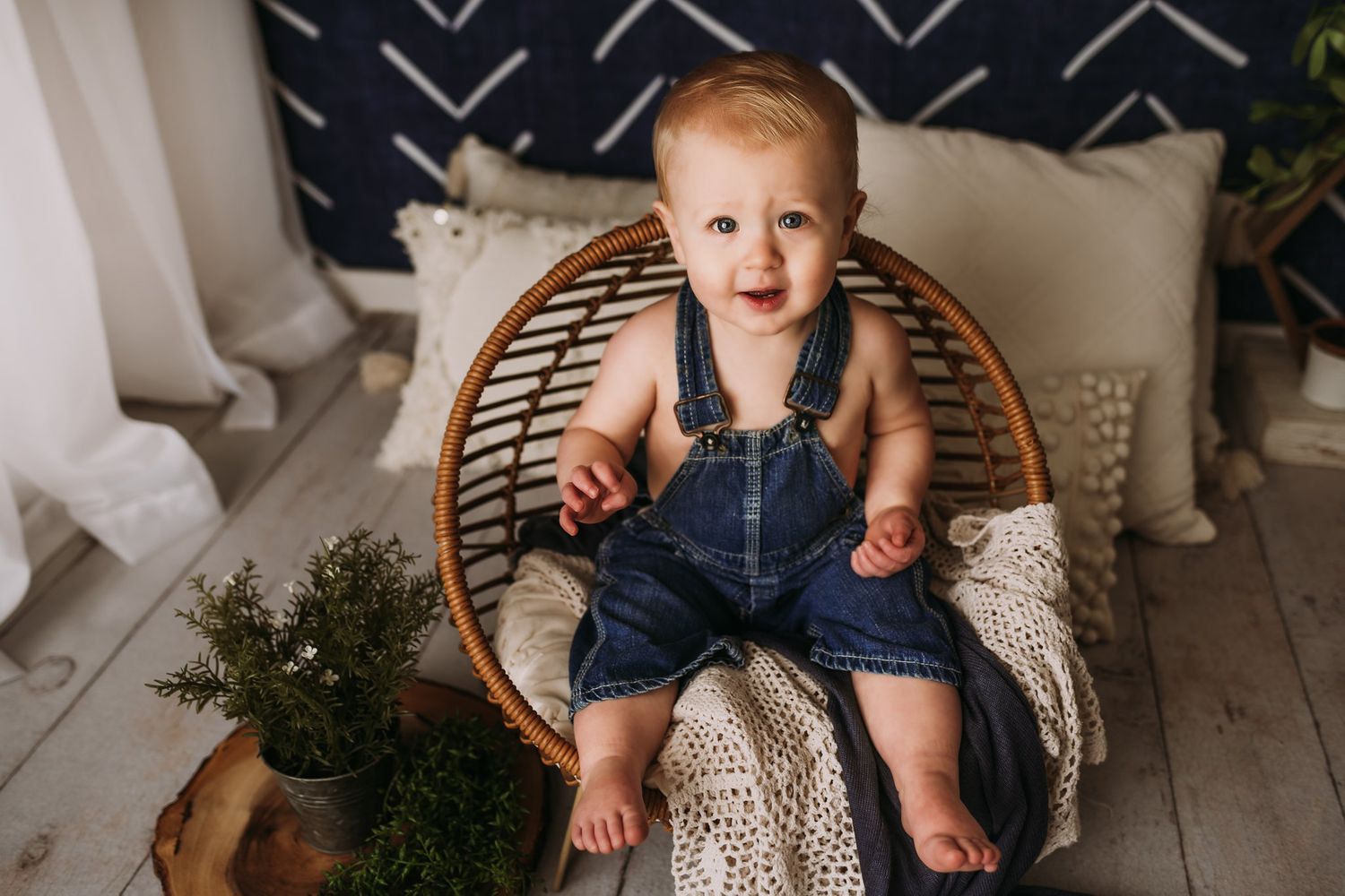 Happy baby wearing denim overalls sits in round wicker chair with decorative backdrop.