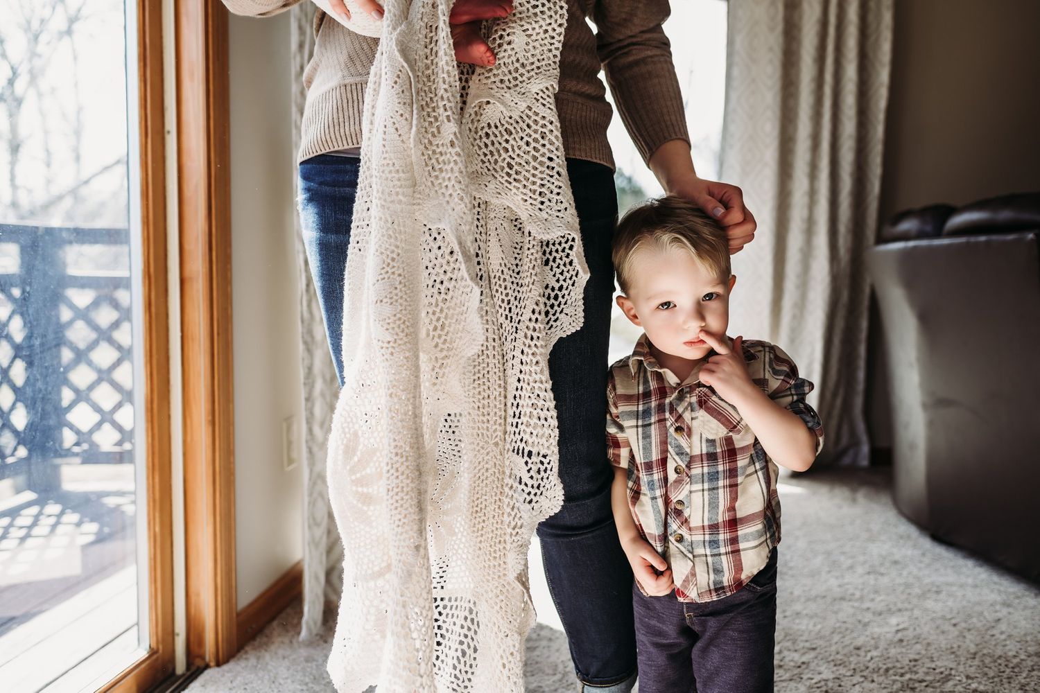 Small child in plaid shirt plays near window with cream lace blanket hanging nearby.