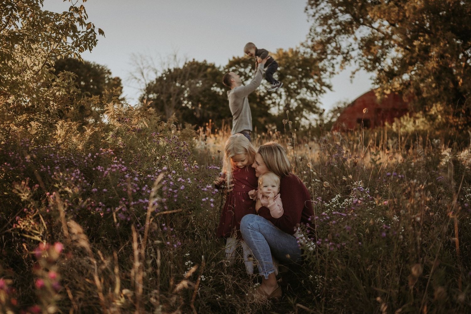 A family enjoying a playful moment together in a wildflower meadow with a red barn in the background.