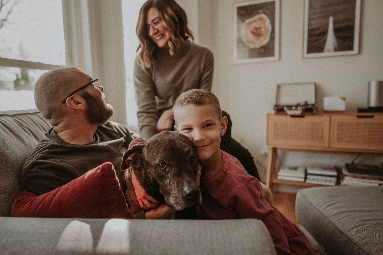 Family cuddles on a gray couch with their dark-colored dog in a modern living room.
