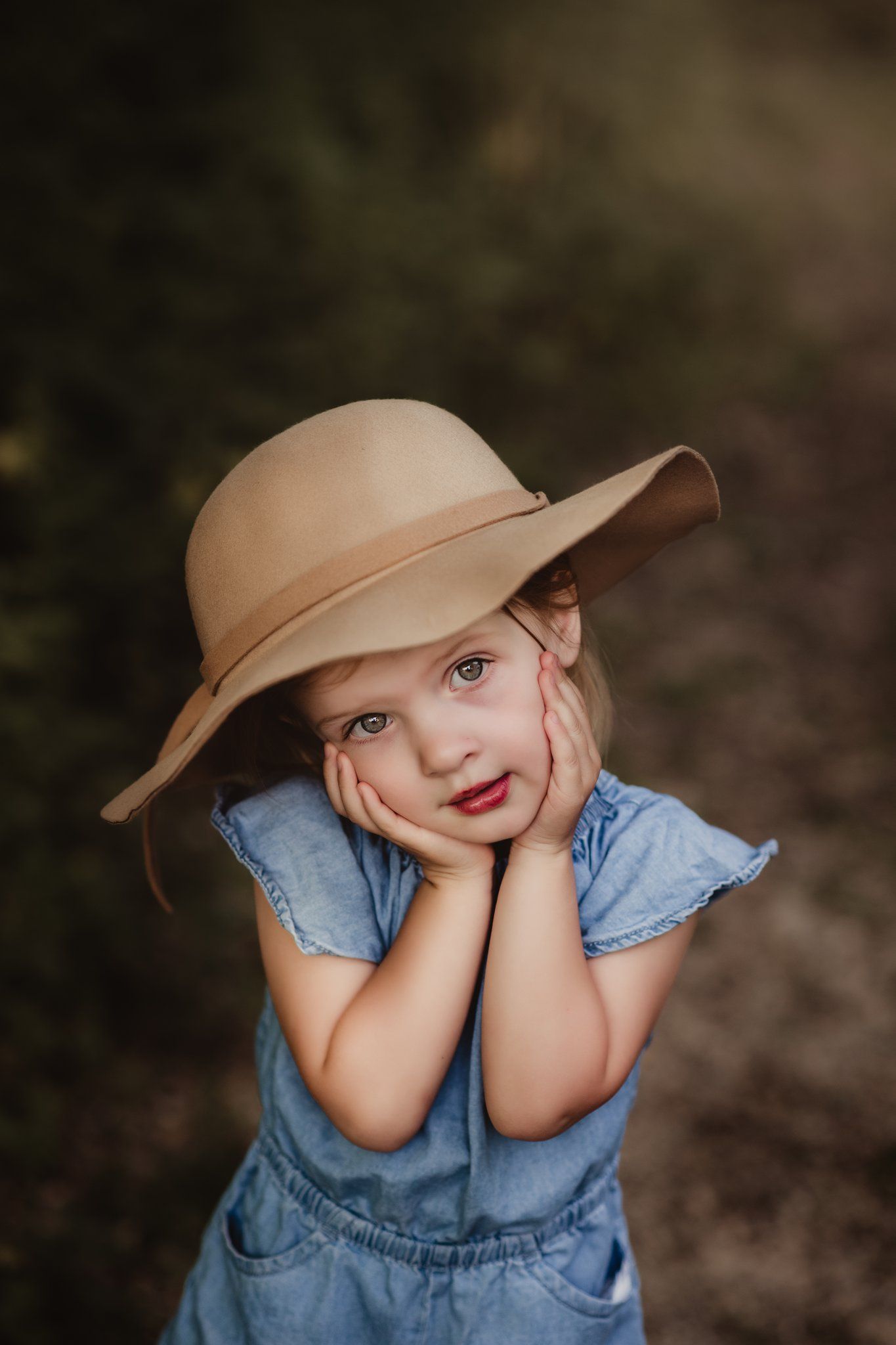 A whimsical portrait in a denim romper and wide-brimmed hat against a dark forest background.
