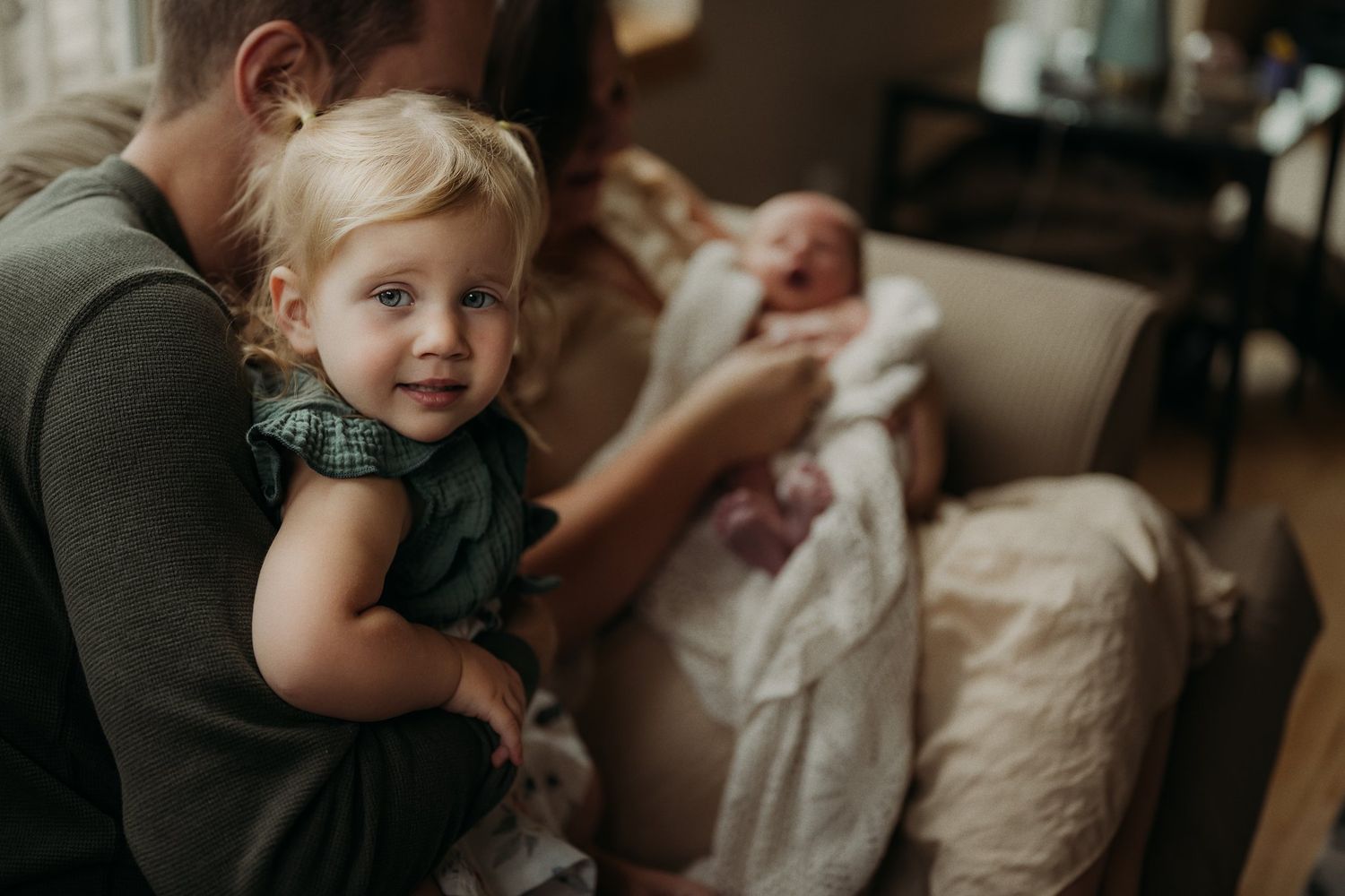 A toddler in a dark dress smiles while sitting with others on a couch.